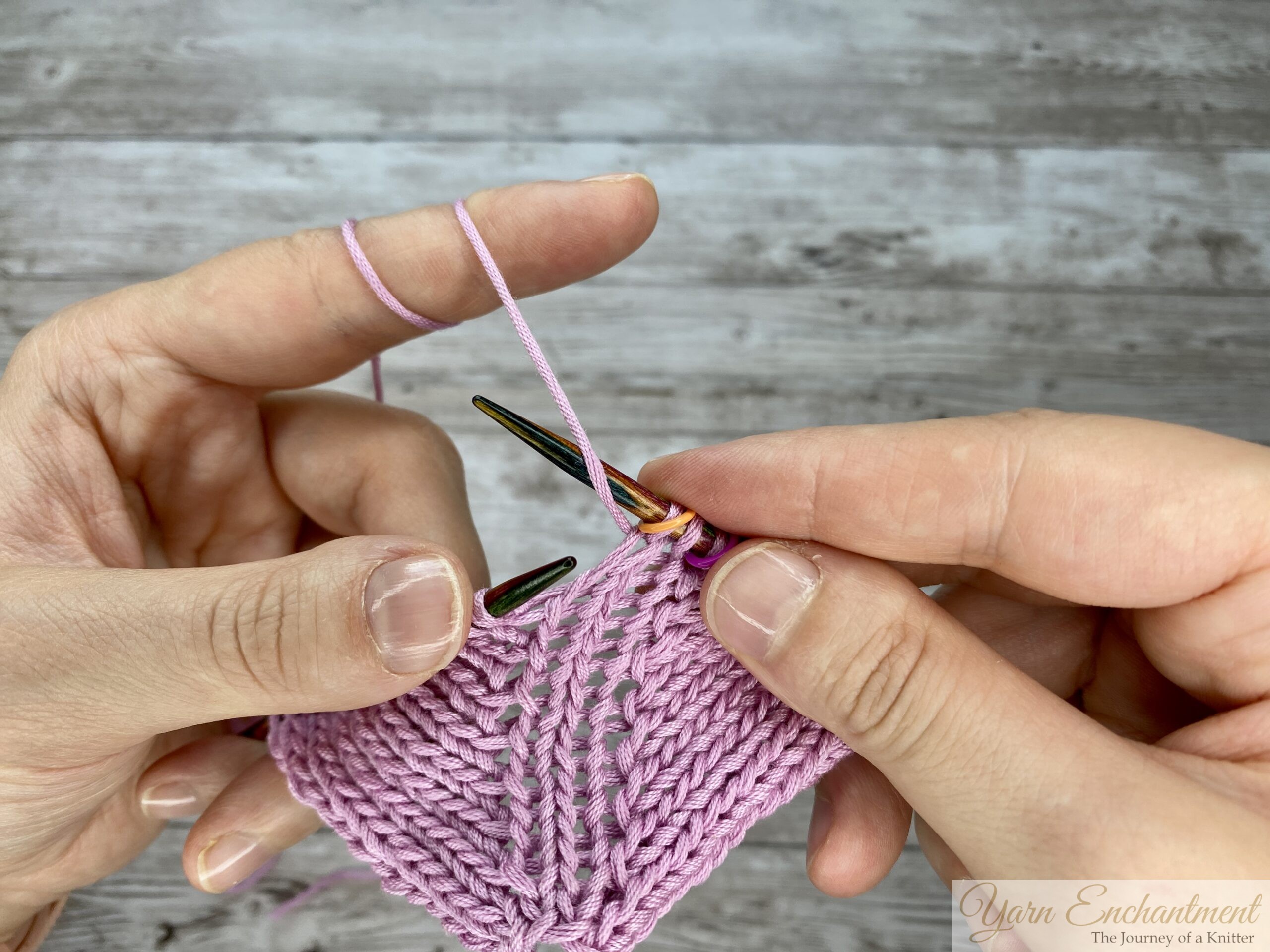 Close-up of hands purling through the back loop (PTBL) on a circular knitting needle with light pink yarn, demonstrating a yarn-over increase technique to prevent laddering and holes in purl stitch fabric.