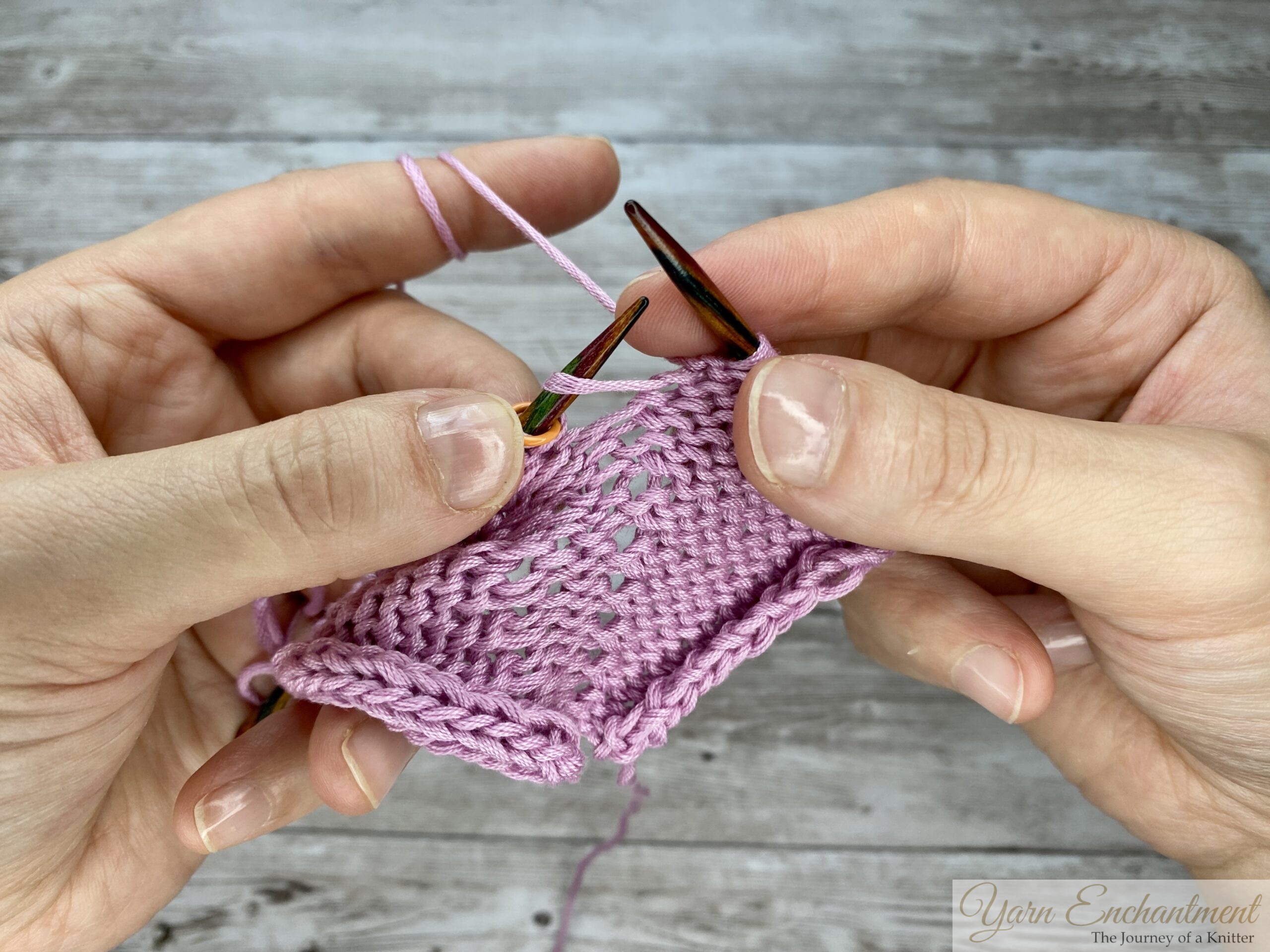 Close-up of hands purling through the back loop (PTBL) on a circular knitting needle with light pink yarn, demonstrating a yarn-over increase technique to prevent laddering and holes in purl stitch fabric