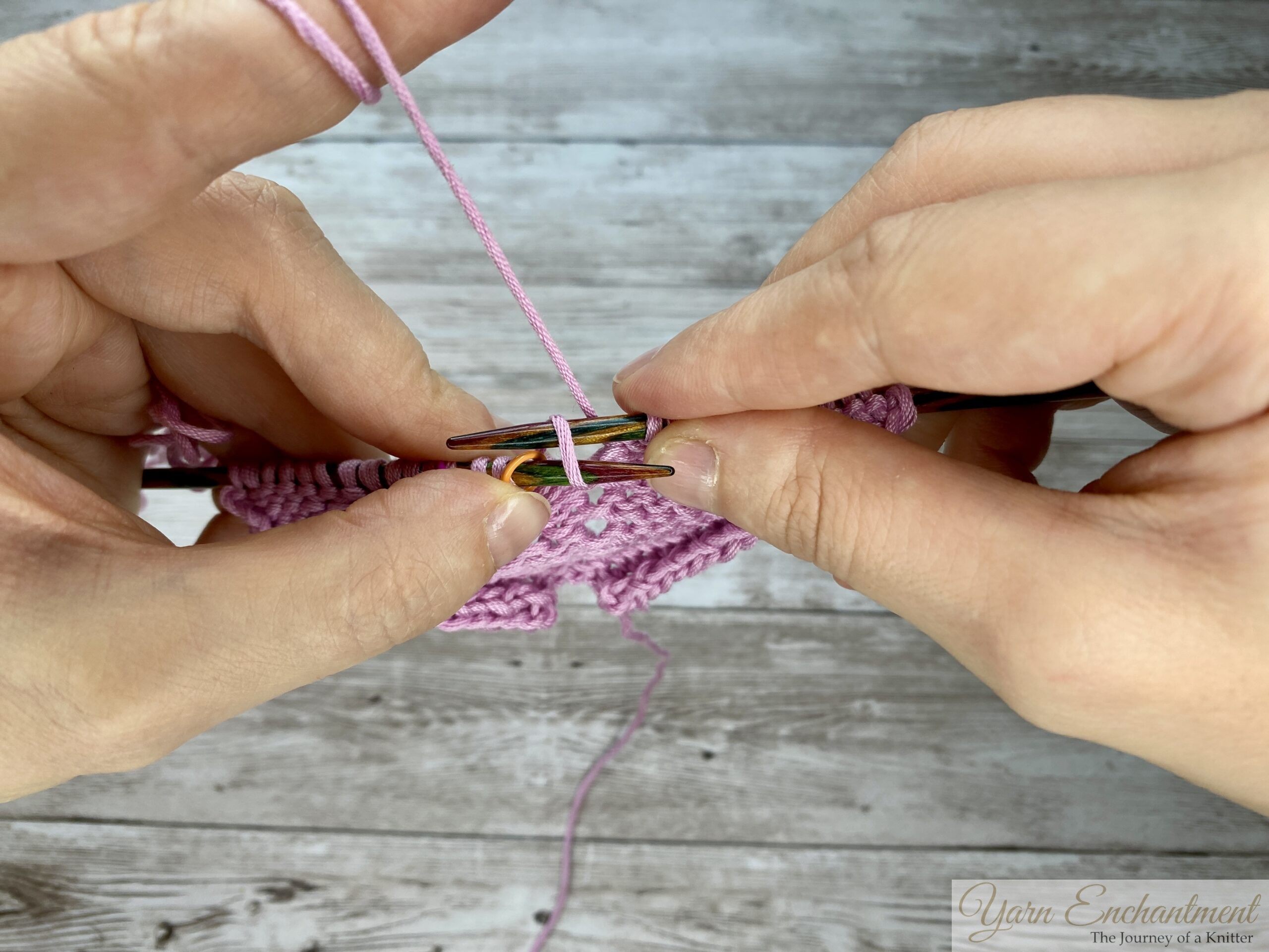 Close-up of hands purling through the back loop (PTBL) on a circular knitting needle with light pink yarn, demonstrating a yarn-over increase technique to prevent laddering and holes in purl stitch fabric