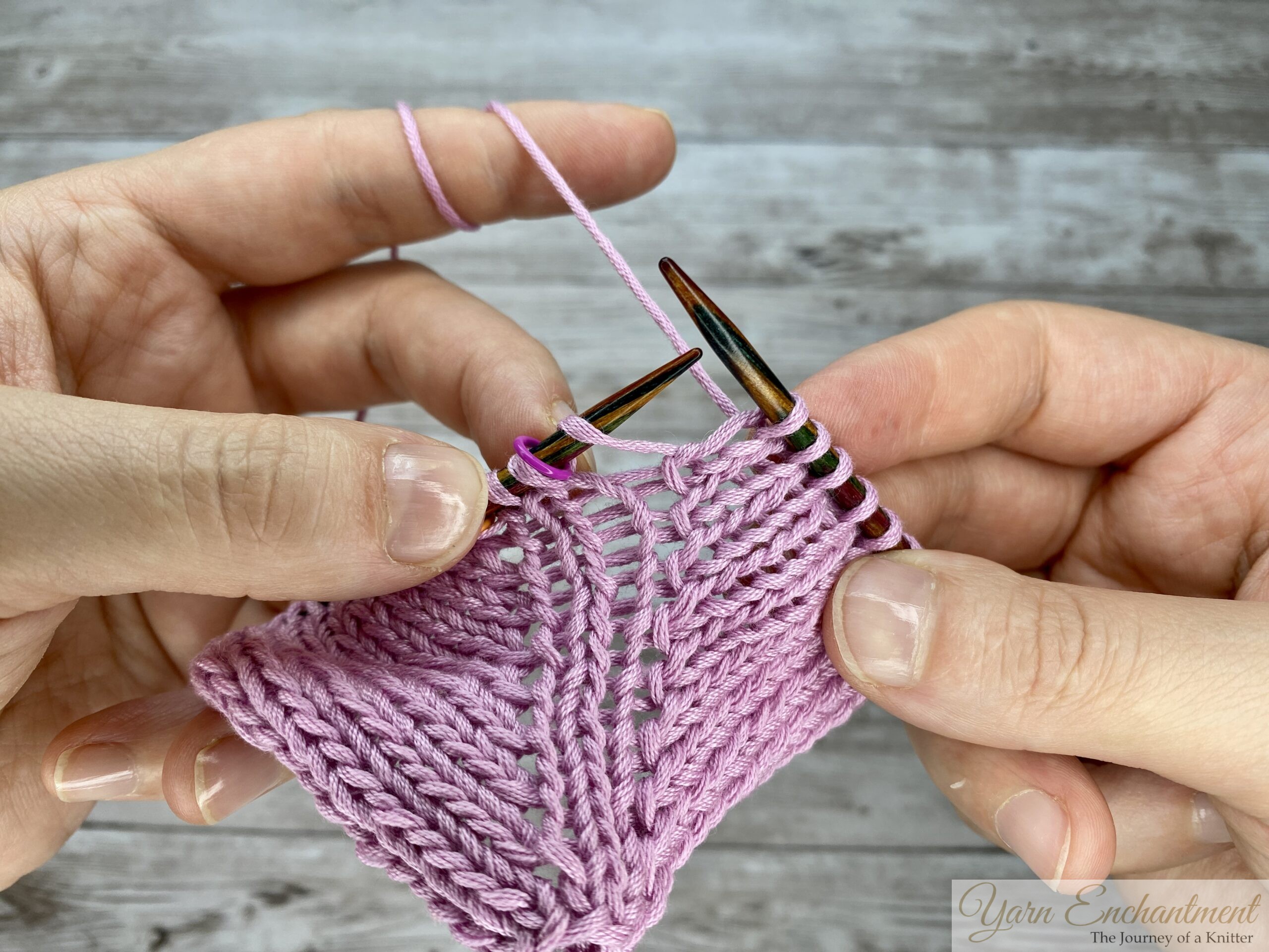 Hands holding pink yarn and circular knitting needles, preparing to execute a yarn-over increase at a stitch marker while working a project in the round.