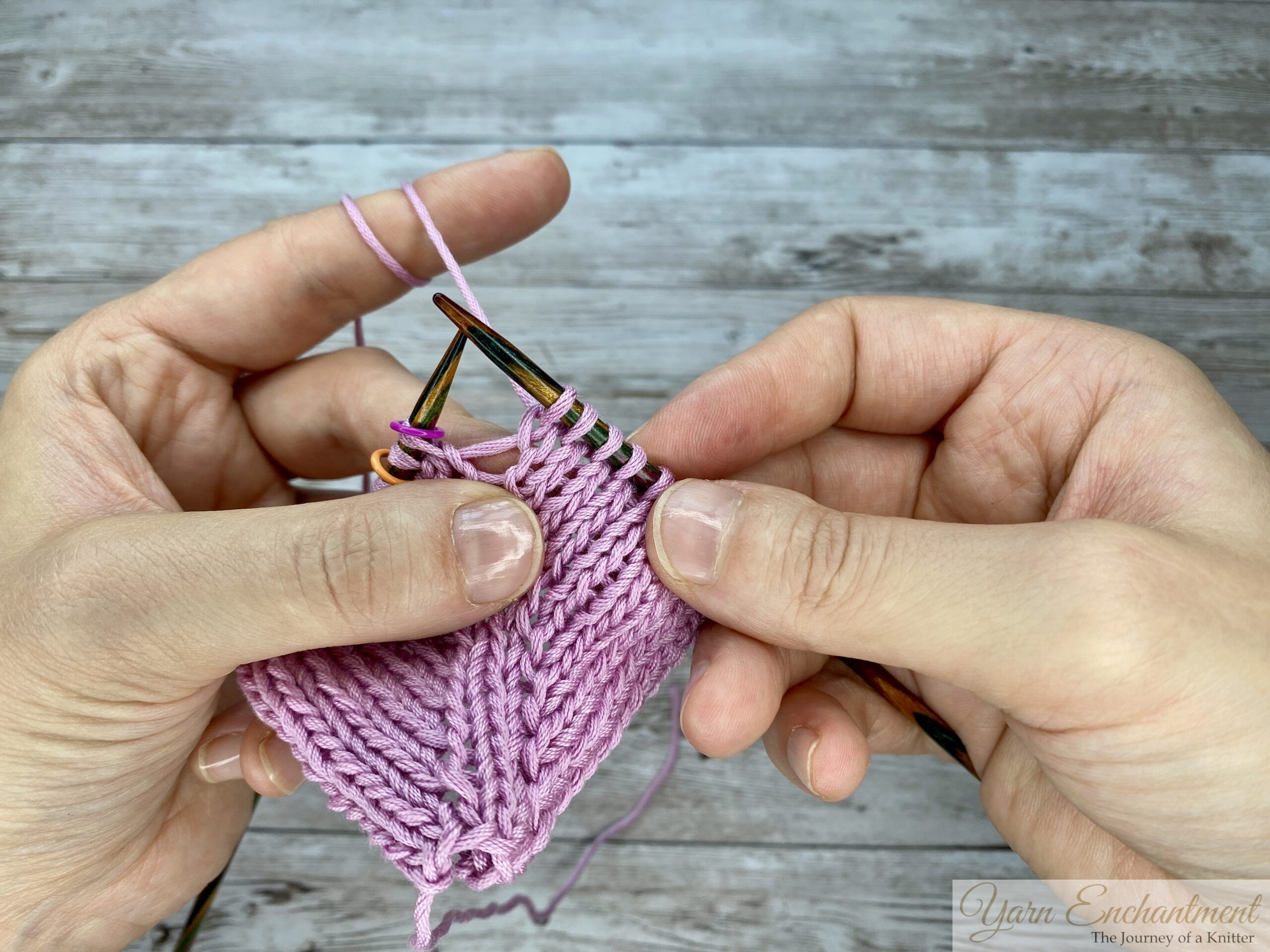 A close-up view of the completed yarn-over loop resting on the right needle, showing how the increase creates an open space in the pink knitted fabric during in-the-round knitting.