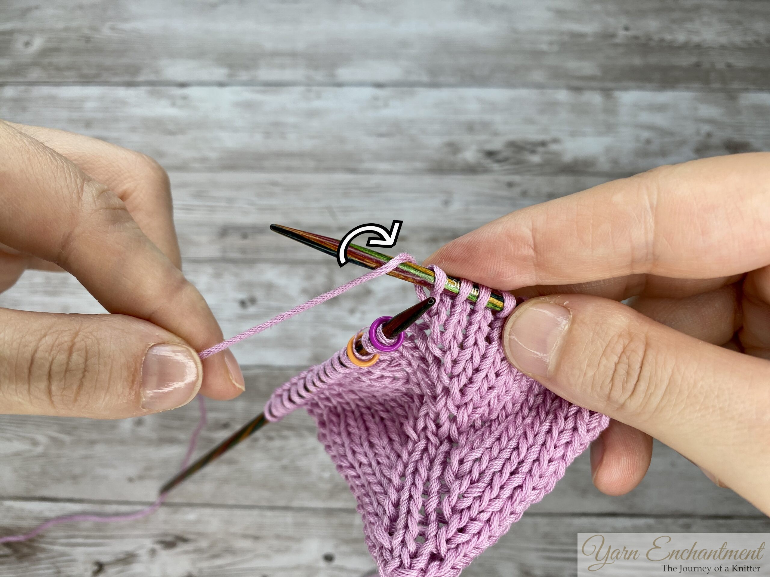 Knitting hands demonstrating a directional M1R increase in the round, with a curved arrow overlay showing how to wrap the pink yarn forward and over the needle to create a right-leaning eyelet.