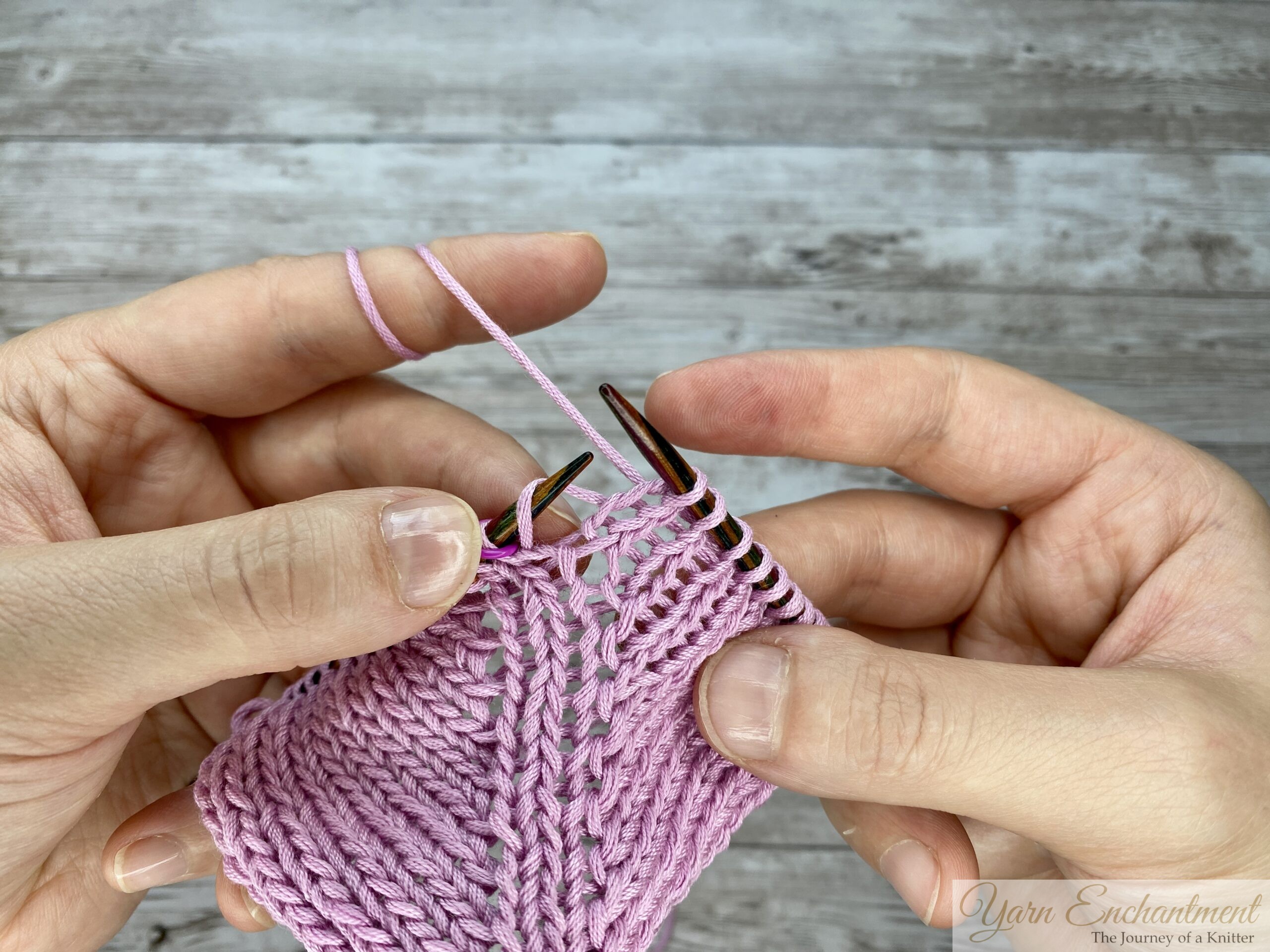 Circular knitting needles holding pink yarn, with the right needle positioned to knit the stitch immediately following a completed directional M1R yarn-over increase.