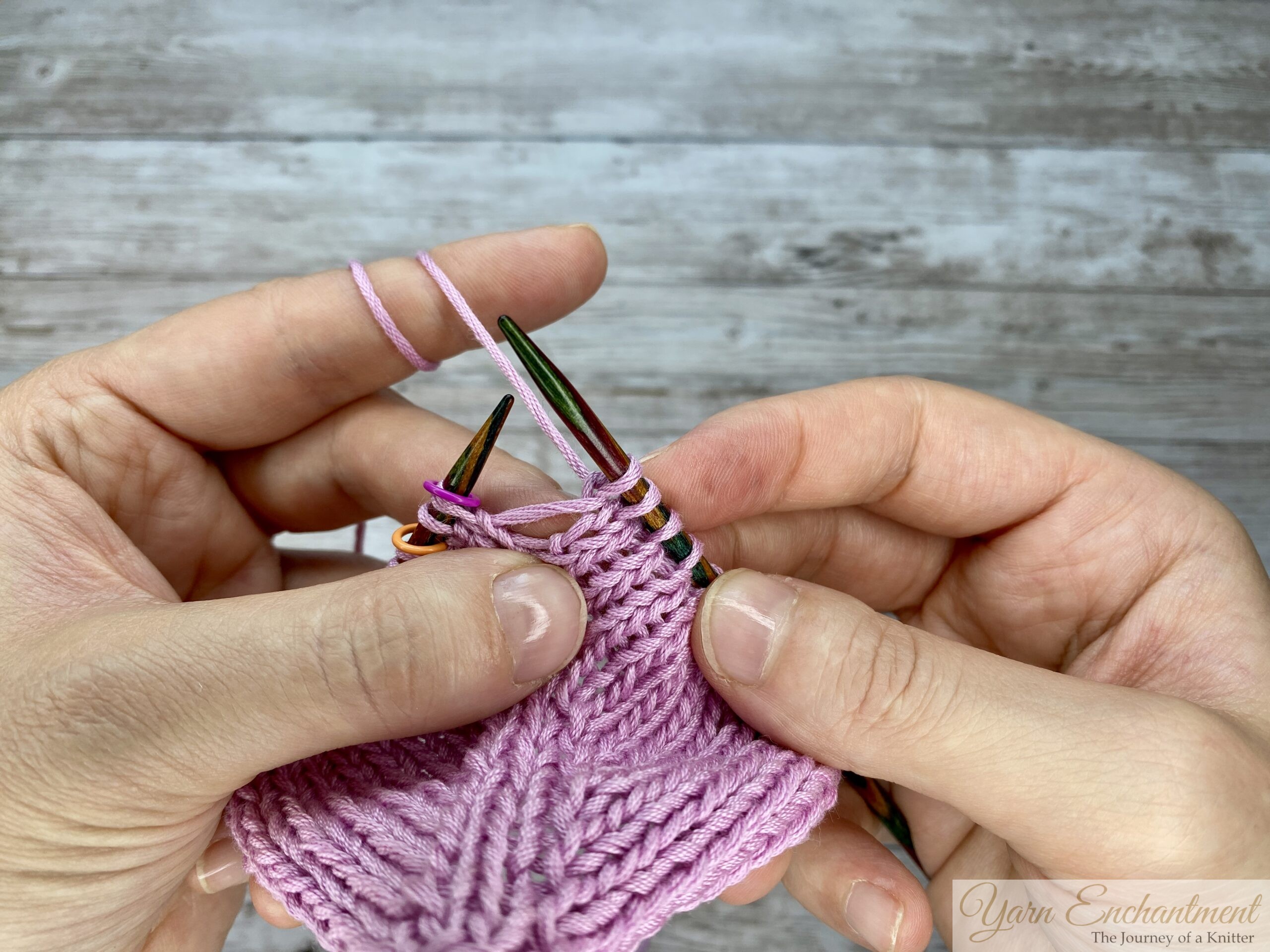 A close-up view of the newly knitted stitch on the right needle, sitting tightly beside the directional M1R increase, ensuring the open eyelet hole remains perfectly framed in the pink garter stitch fabric.
