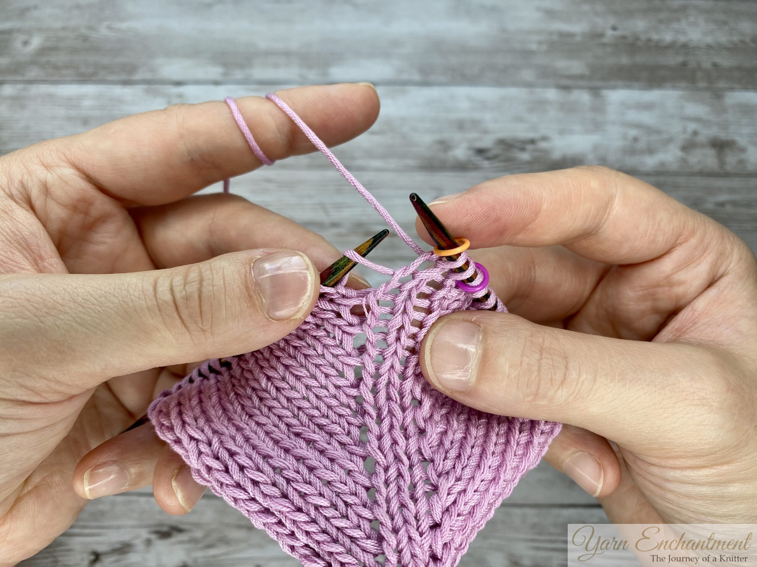 Hands holding circular knitting needles and pink yarn, positioning the right needle to knit the stitch directly after a directional yarn-over increase while working in the round.