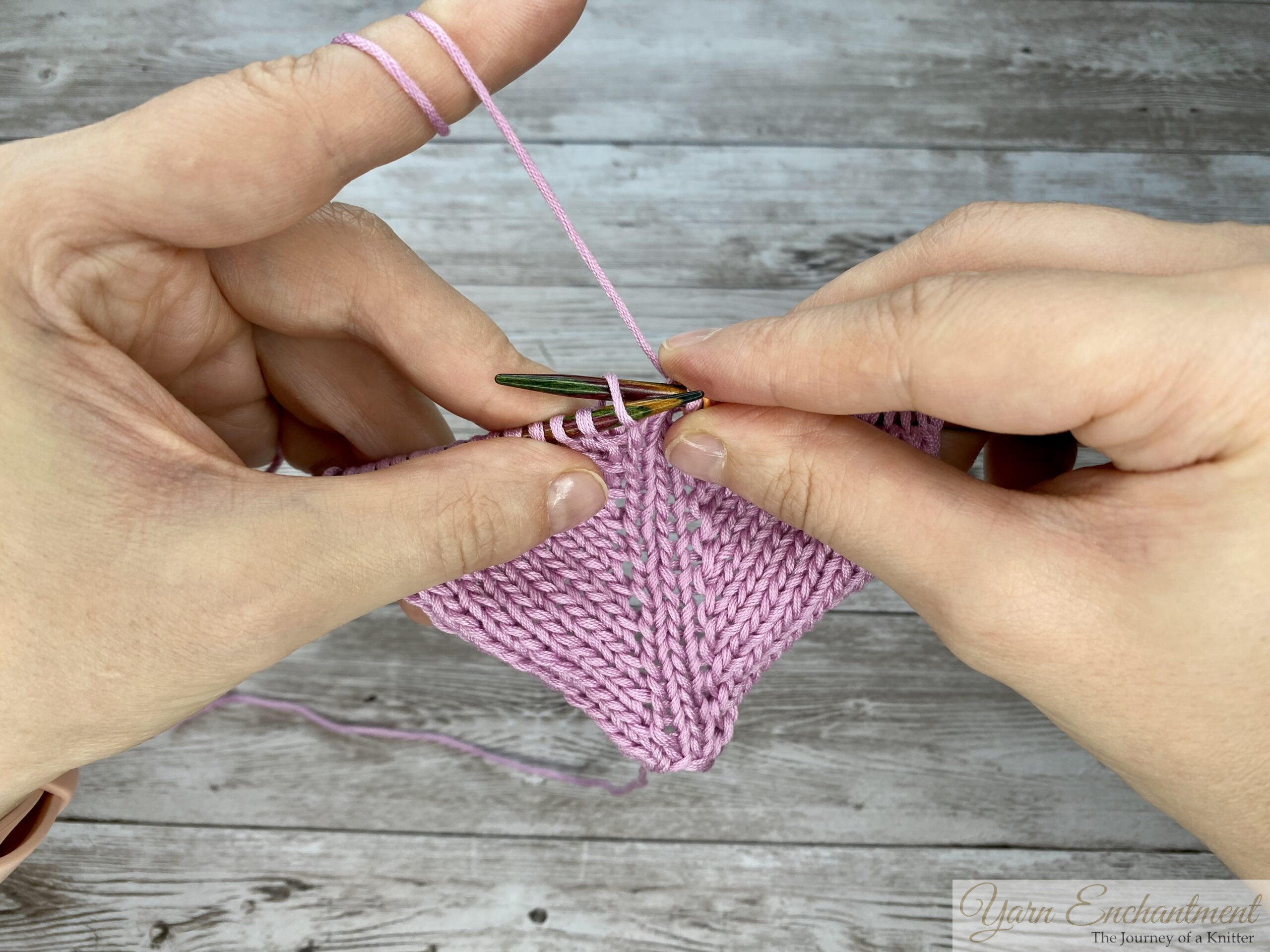Wrapping the pink working yarn to complete the knit stitch following the directional YO, maintaining an open eyelet loop on the right-hand needle.