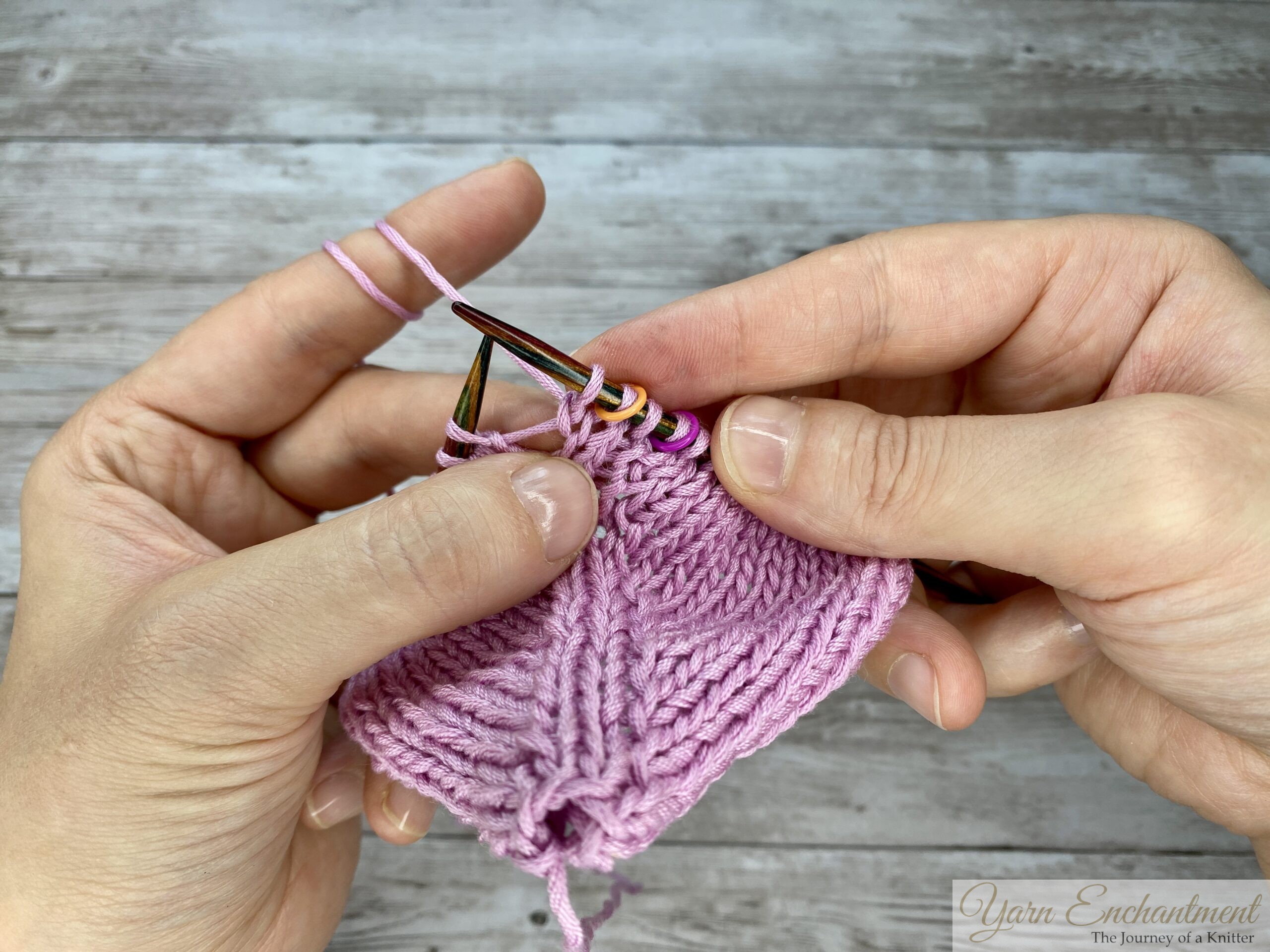The knit stitch after the yarn-over is completed and transferred to the right needle, revealing the open left-leaning eyelet hole created by the directional increase on this in-the-round pink project.