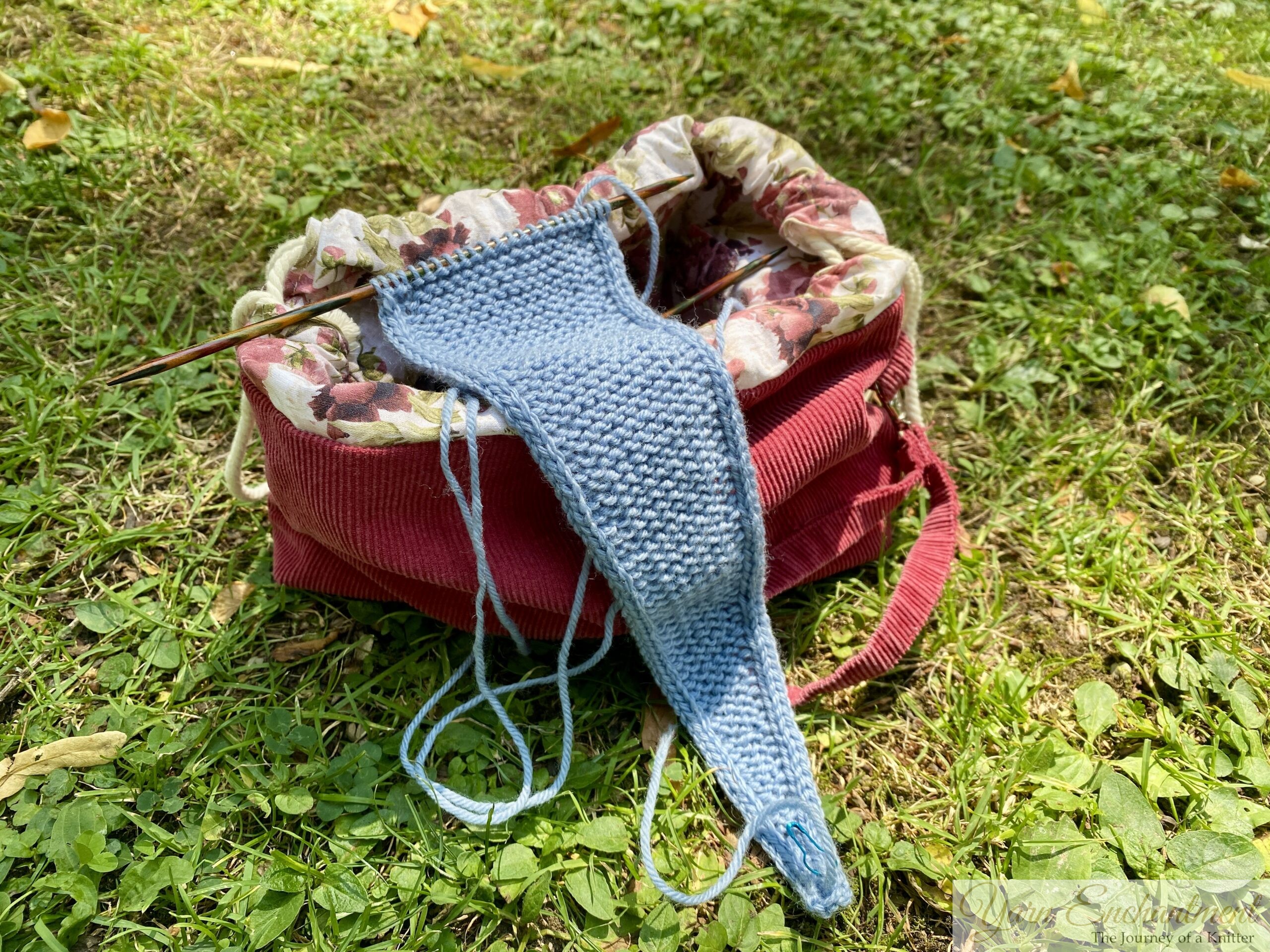 A work-in-progress light blue knitted scarf on wooden needles resting inside a handmade project bag. The bag is made from dark red corduroy with a floral fabric lining, sitting outdoors on green grass in the sunlight.