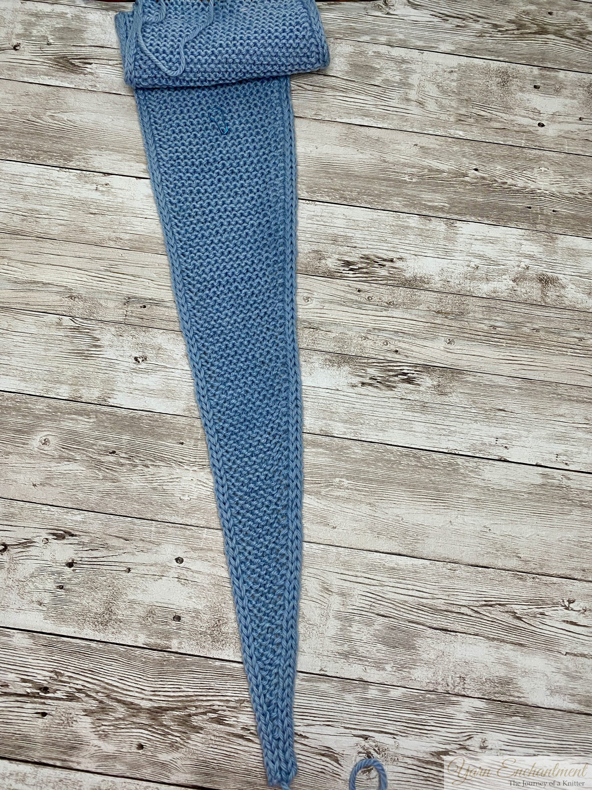 A partially finished light blue Sophie scarf laid flat on a weathered grey wooden surface. The scarf is shaped as a long, narrowing triangle, knitted in a garter stitch pattern with prominent horizontal ridges. Both long edges feature a distinct, thick braided i-cord border. The wider, top end is slightly folded over, while the narrow tip tapers down to a point at the bottom, with a small loop of yarn resting nearby on the wood.
