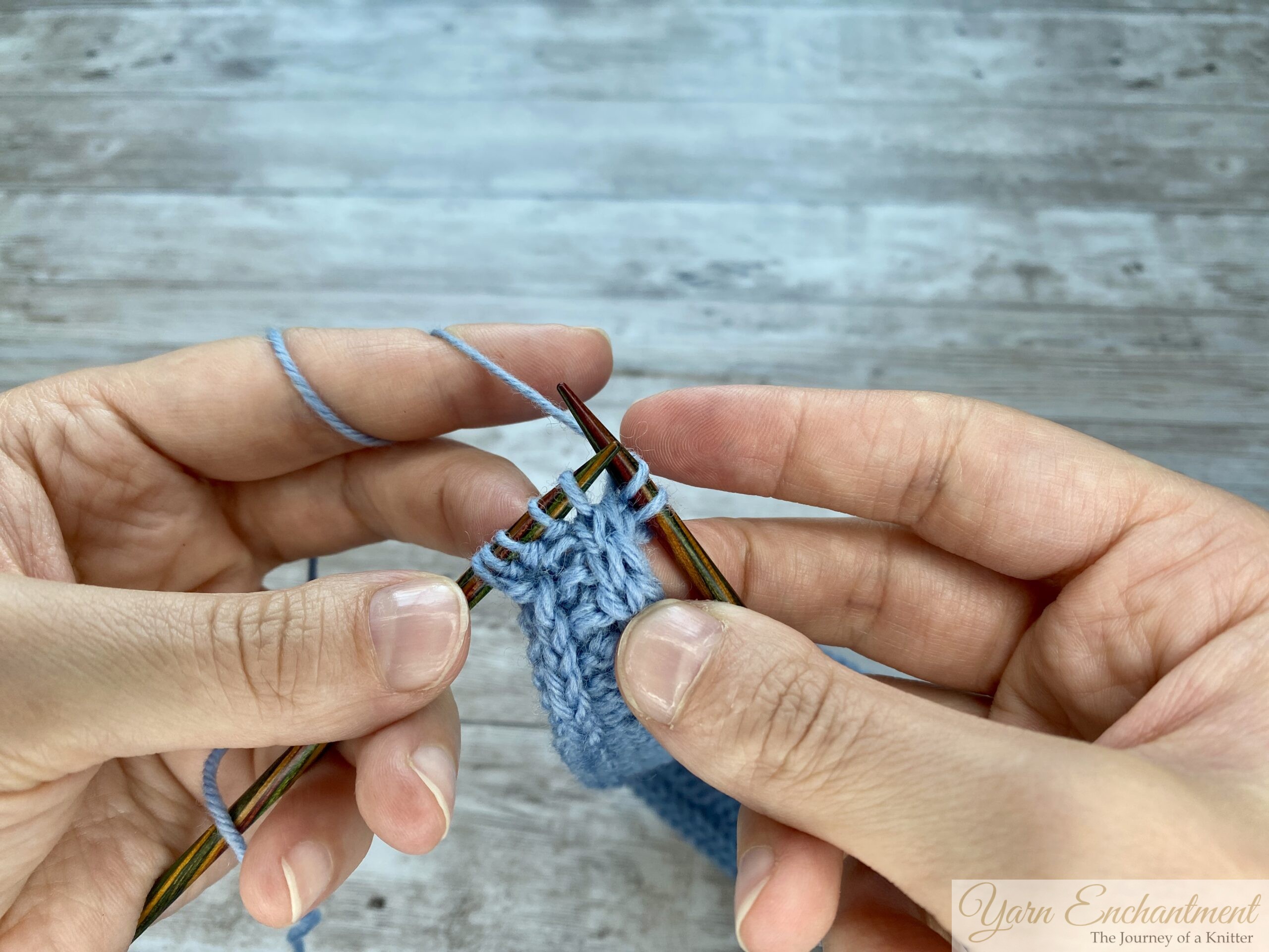 Hands holding wooden knitting needles working a decrease on a light blue Sophie scarf, showing the step-by-step motion of passing the working yarn over and pulling through the loops.
