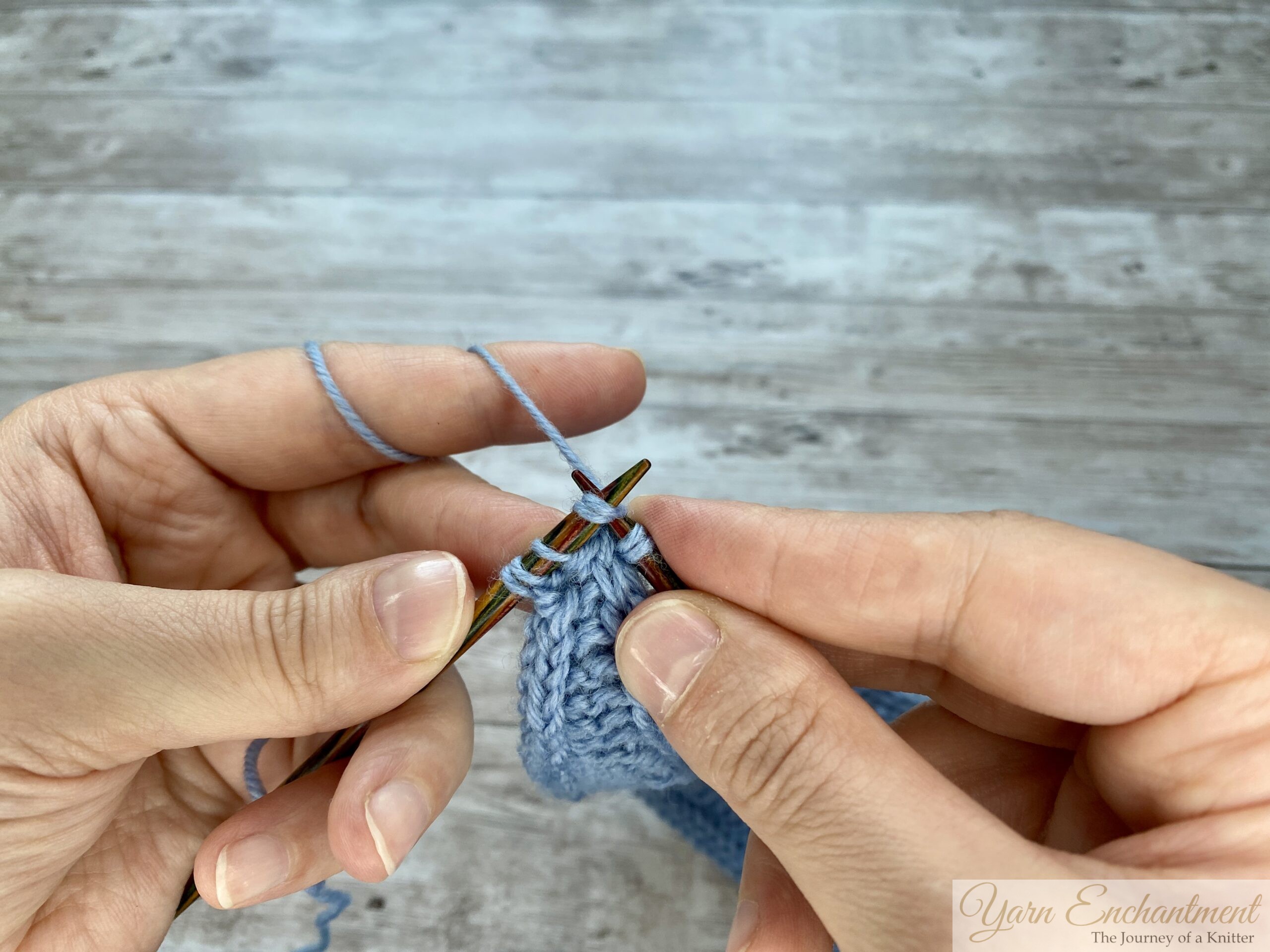 Hands holding wooden knitting needles working a decrease on a light blue Sophie scarf, showing the step-by-step motion of passing the working yarn over and pulling through the loops.