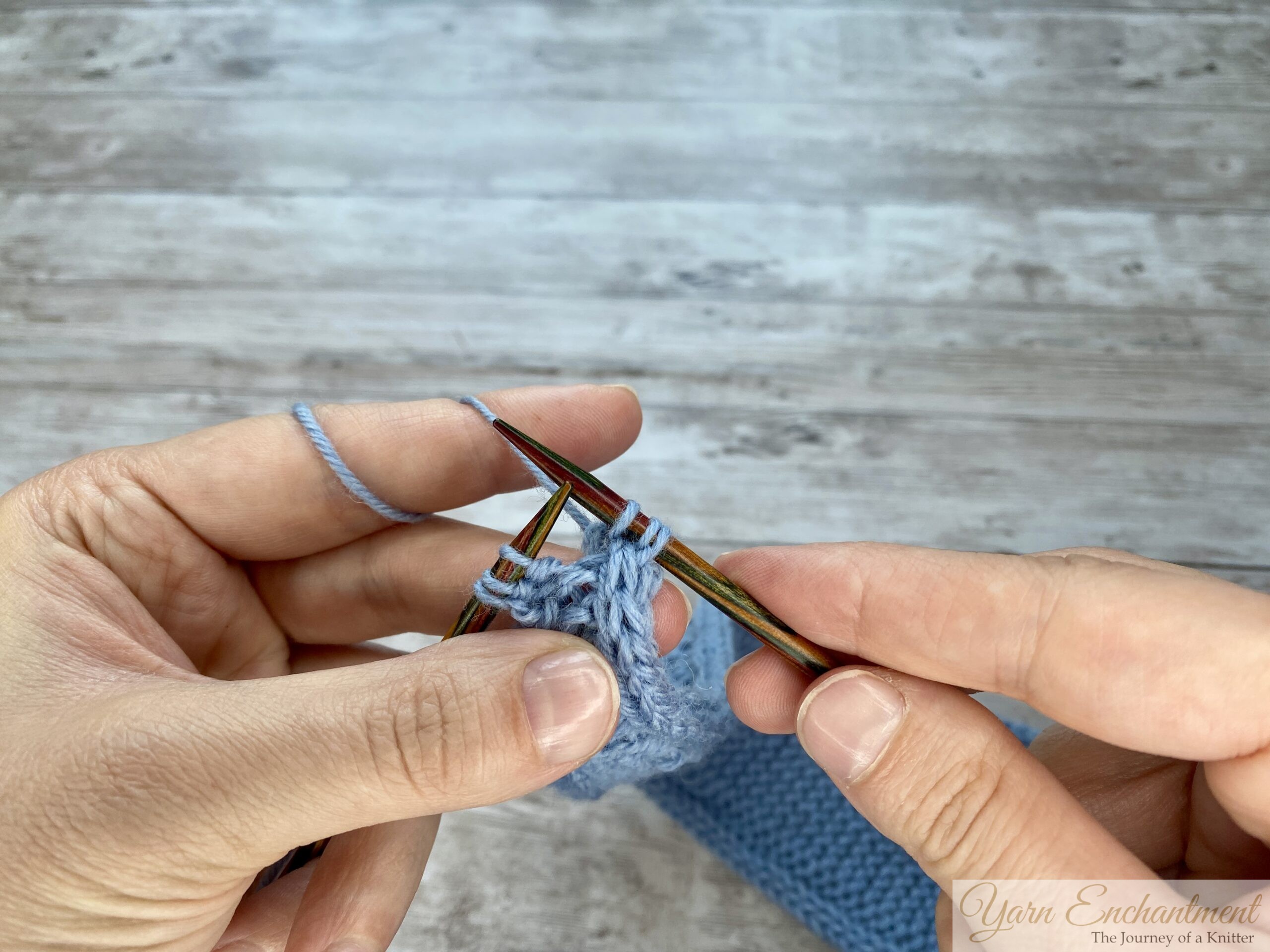 Hands holding wooden knitting needles working a decrease on a light blue Sophie scarf, showing the step-by-step motion of passing the working yarn over and pulling through the loops.
