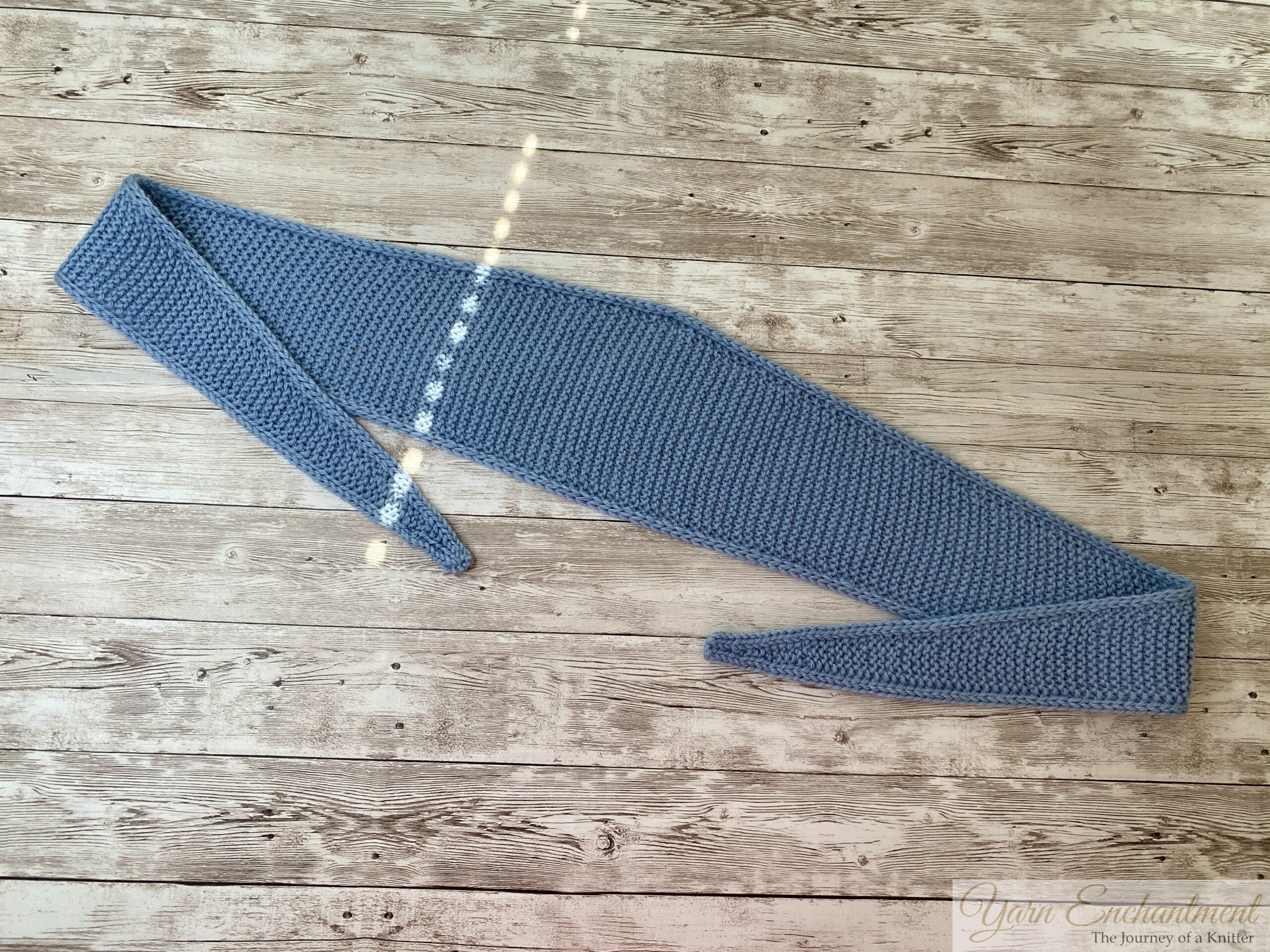 A finished light blue Sophie scarf laid flat and stretched out across a rustic wooden surface. The scarf is shaped like a long, curved band that is wide in the middle and tapers to a point at both ends.