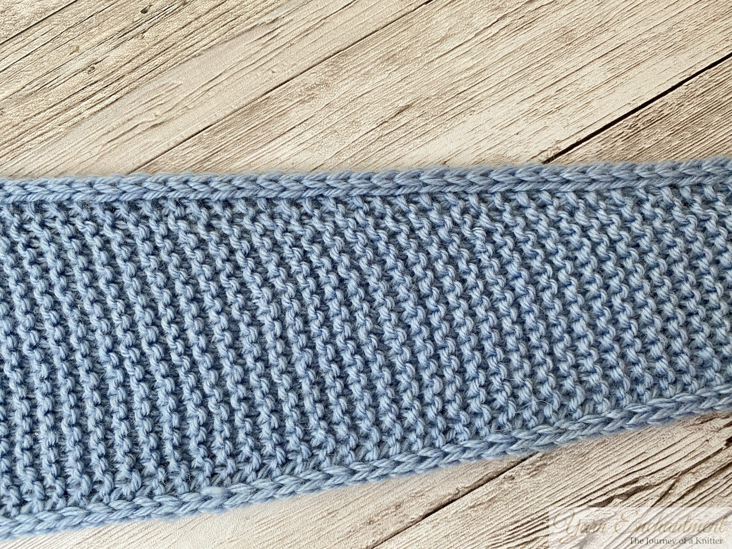 A close-up, overhead shot of a light blue knitted Sophie scarf laid flat on whitewashed wood, showing the neat horizontal ridges of the garter stitch and the braided edge.