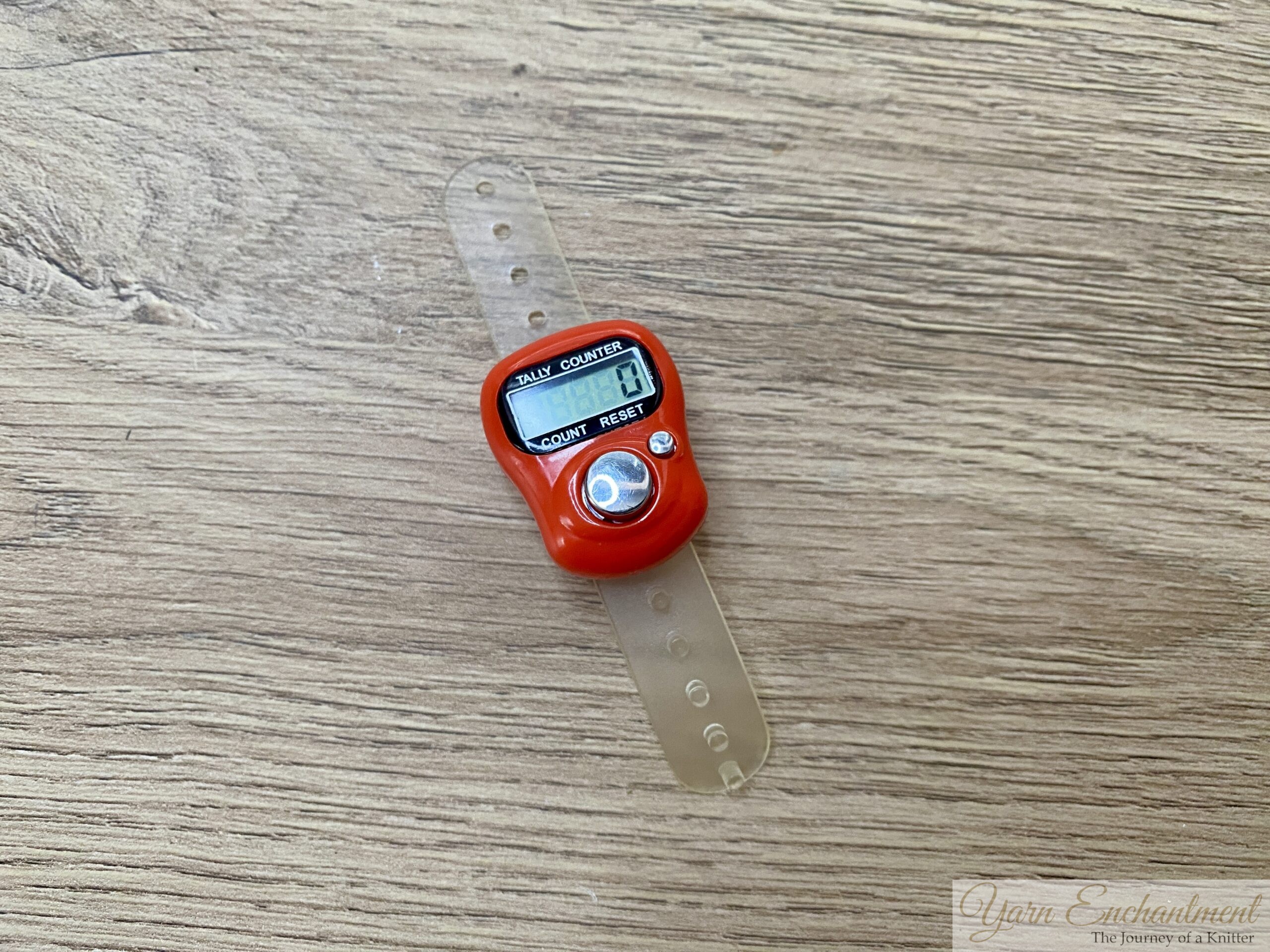 A small, bright red digital tally counter with a clear plastic finger strap, resting on a light wooden surface. The screen displays the number 0.