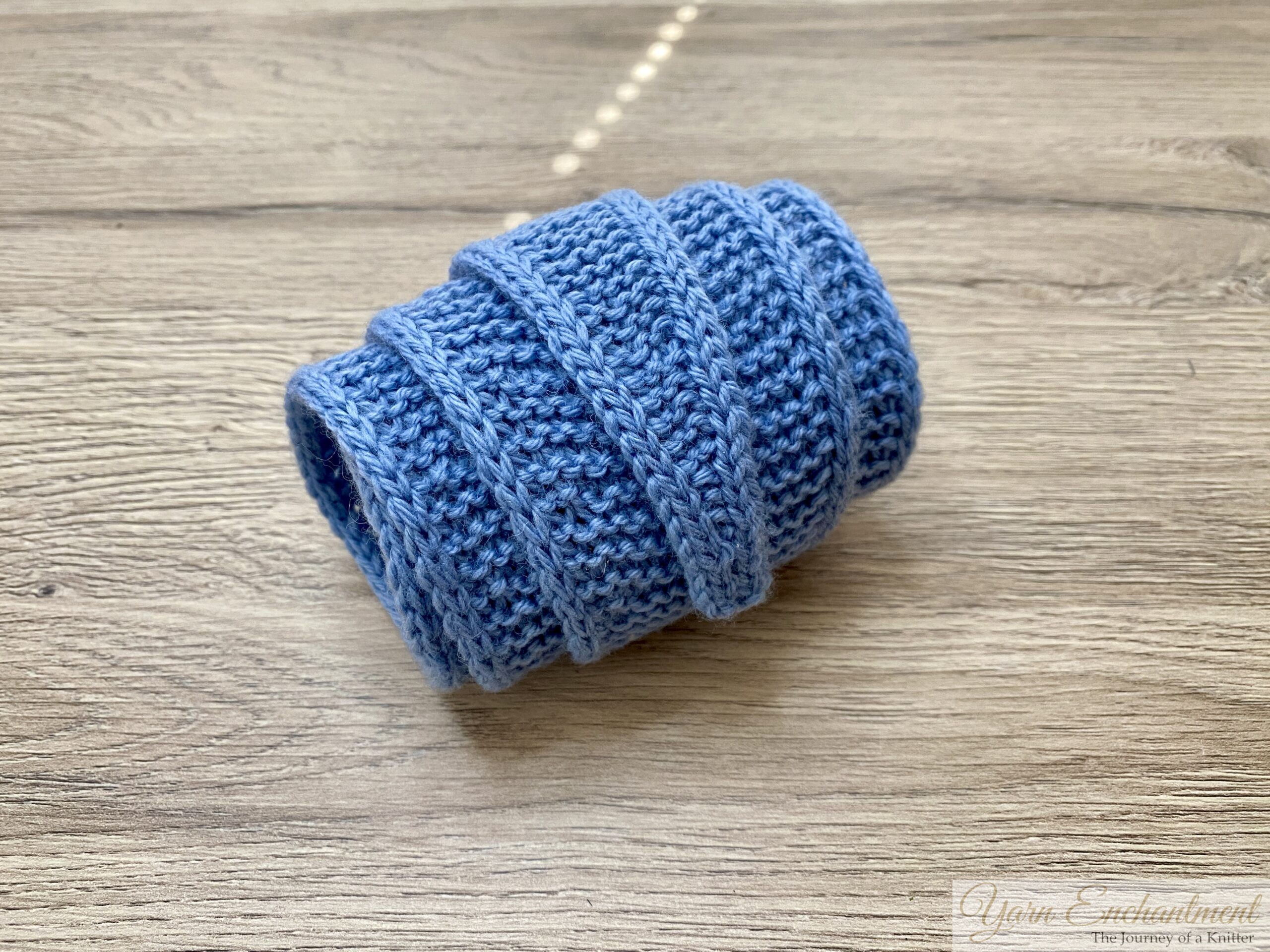 A light blue knitted Sophie scarf rolled tightly into a neat, cylindrical tube, resting on a rustic wooden table, highlighting the bumpy garter stitch texture.