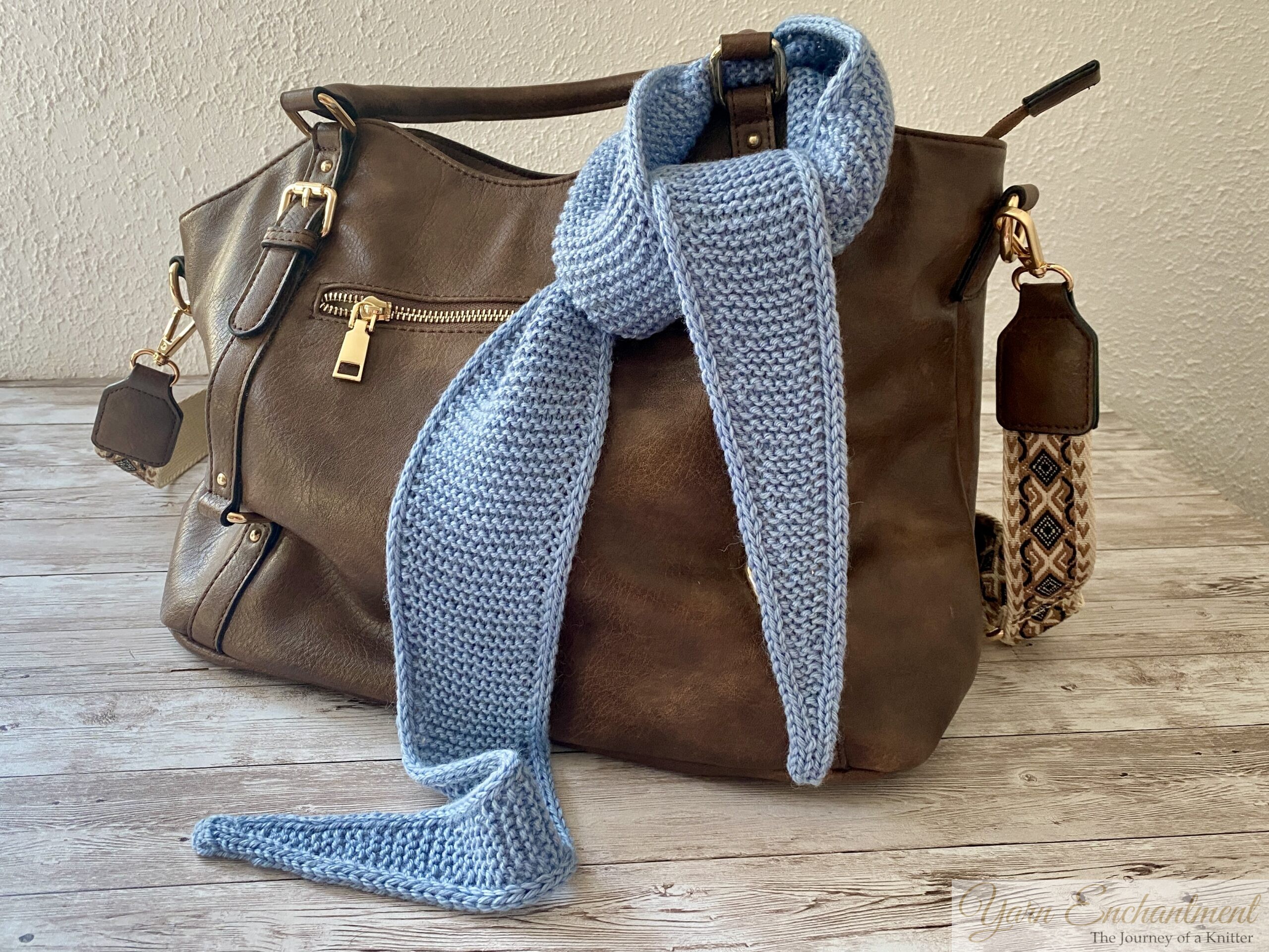 A light blue knitted Sophie scarf draped around the handle of a brown leather handbag on a rustic wooden table.