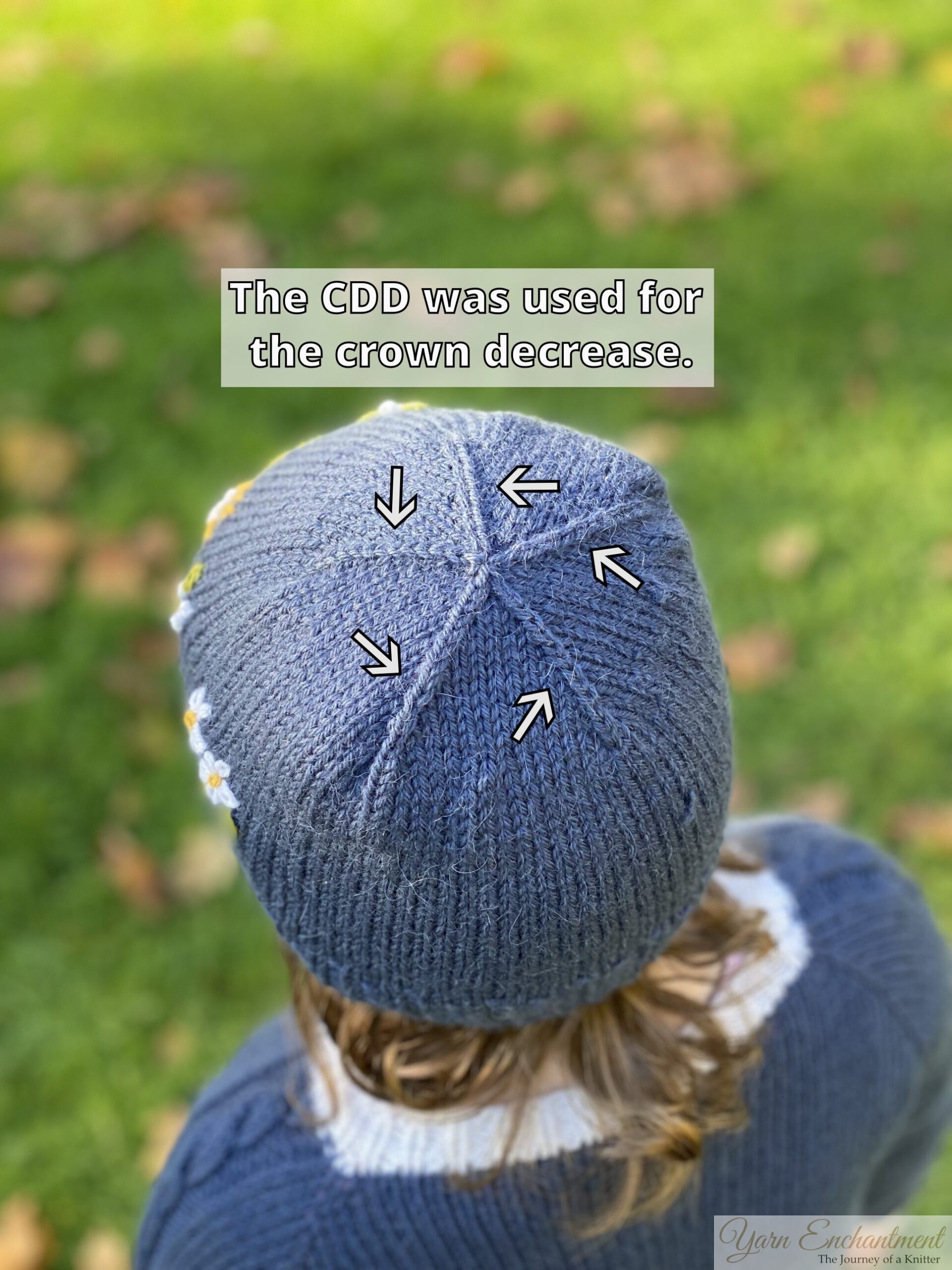A top-down view of a person wearing a blue knit beanie outdoors, with green grass blurred in the background. The crown of the hat features several subtle, raised vertical ridges that meet at a center point, created by decreasing stitches. Five white arrows with black outlines point directly to these ridges. A text box overlay reads, 'The CDD was used for the crown decrease.' The hat has small white daisy motifs on the side and the wearer has curly brown hair.
