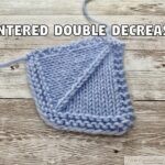 A light blue knitted swatch resting on a wooden surface, demonstrating a 'Centered Double Decrease' in knitting. The stitch pattern shows a distinct, straight diagonal line moving upwards from left to right through the center of the stockinette fabric, bordered by garter stitch edges. Bold white text with a black outline reading 'CENTERED DOUBLE DECREASE' is overlaid at the top of the image.