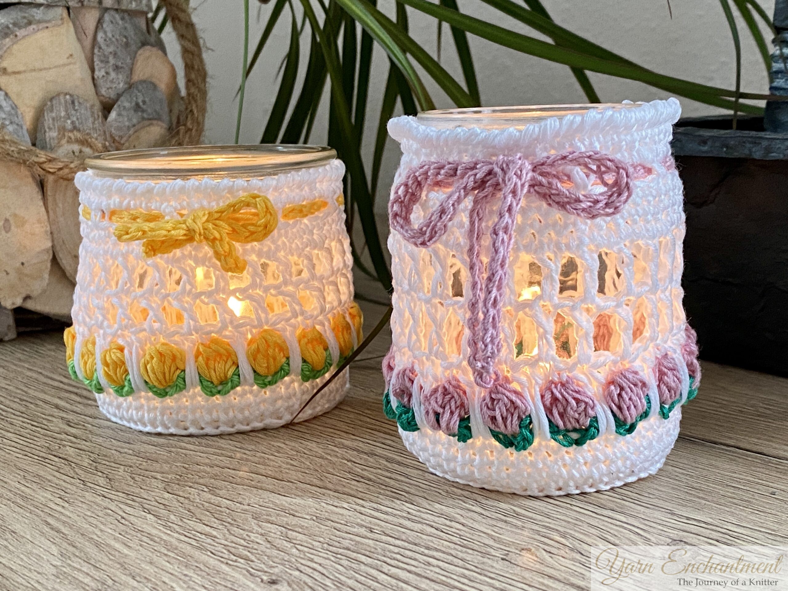 Free crochet pattern for a spring tulip jar cover. Two white mesh crochet cozies adorned with yellow and dusty pink tulip motifs and matching yarn bows, displayed over upcycled glass jars. The second image shows the jars lit from within with a warm LED candle, creating a cozy glow perfect for farmhouse table decor.