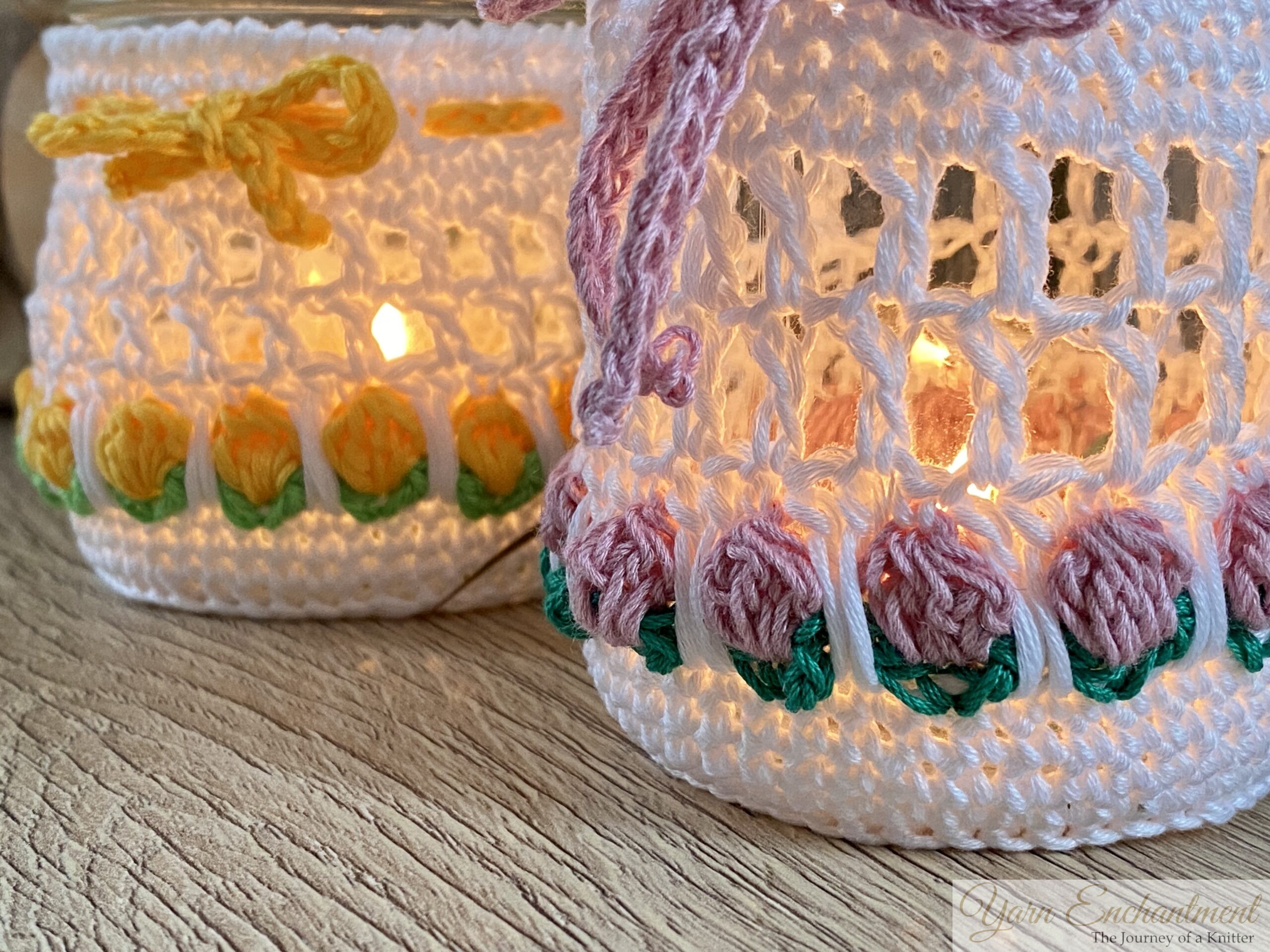 Free crochet pattern for a spring tulip jar cover. Two white mesh crochet cozies adorned with yellow and dusty pink tulip motifs and matching yarn bows, displayed over upcycled glass jars. The second image shows the jars lit from within with a warm LED candle, creating a cozy glow perfect for farmhouse table decor.