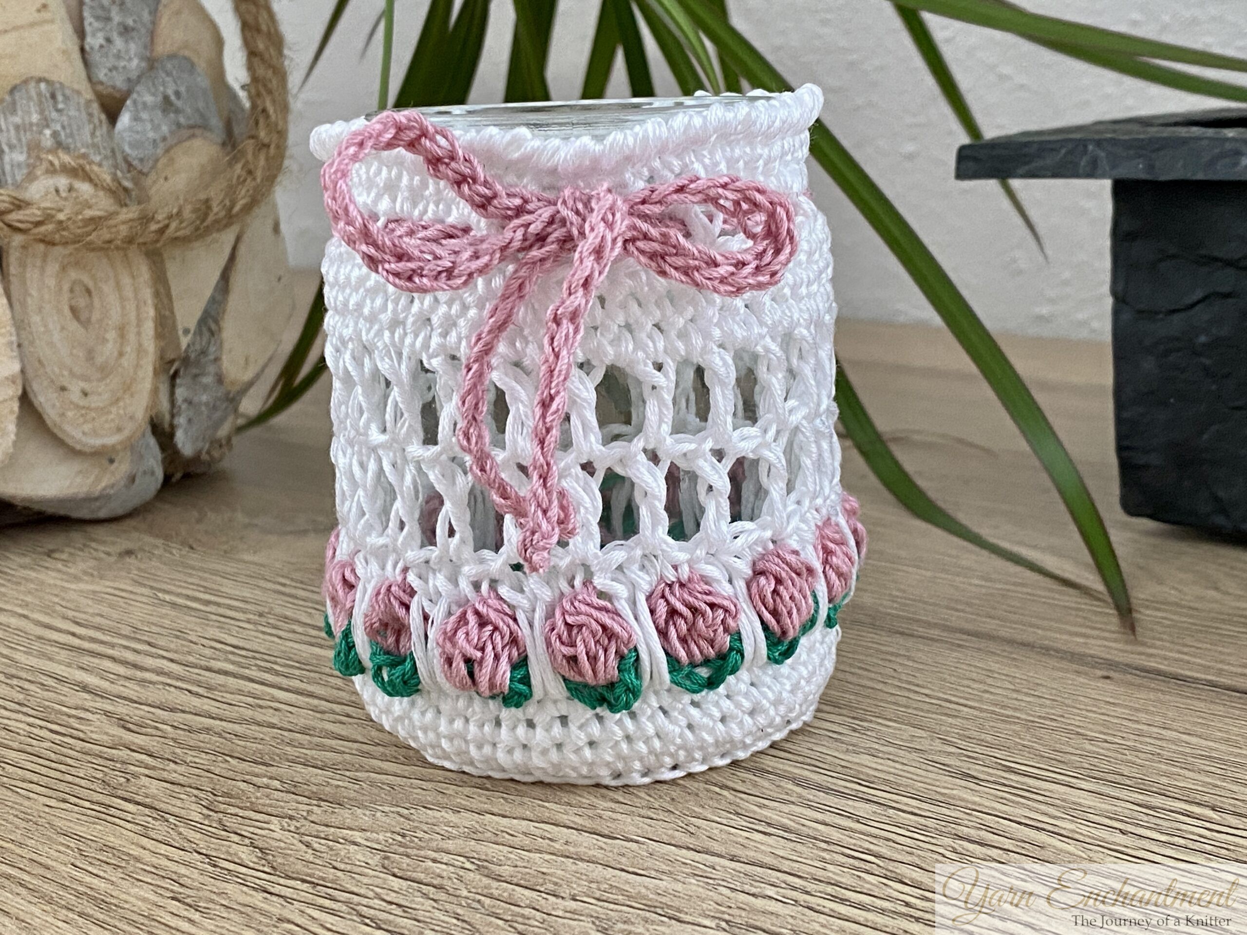 Free crochet pattern for a spring tulip jar cover. Two white mesh crochet cozies adorned with yellow and dusty pink tulip motifs and matching yarn bows, displayed over upcycled glass jars. The second image shows the jars lit from within with a warm LED candle, creating a cozy glow perfect for farmhouse table decor.