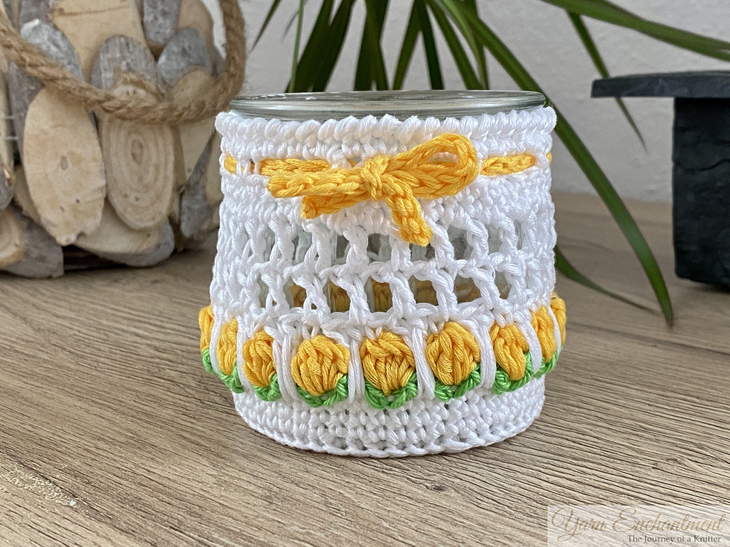 Free crochet pattern for a spring tulip jar cover. Two white mesh crochet cozies adorned with yellow and dusty pink tulip motifs and matching yarn bows, displayed over upcycled glass jars. The second image shows the jars lit from within with a warm LED candle, creating a cozy glow perfect for farmhouse table decor.