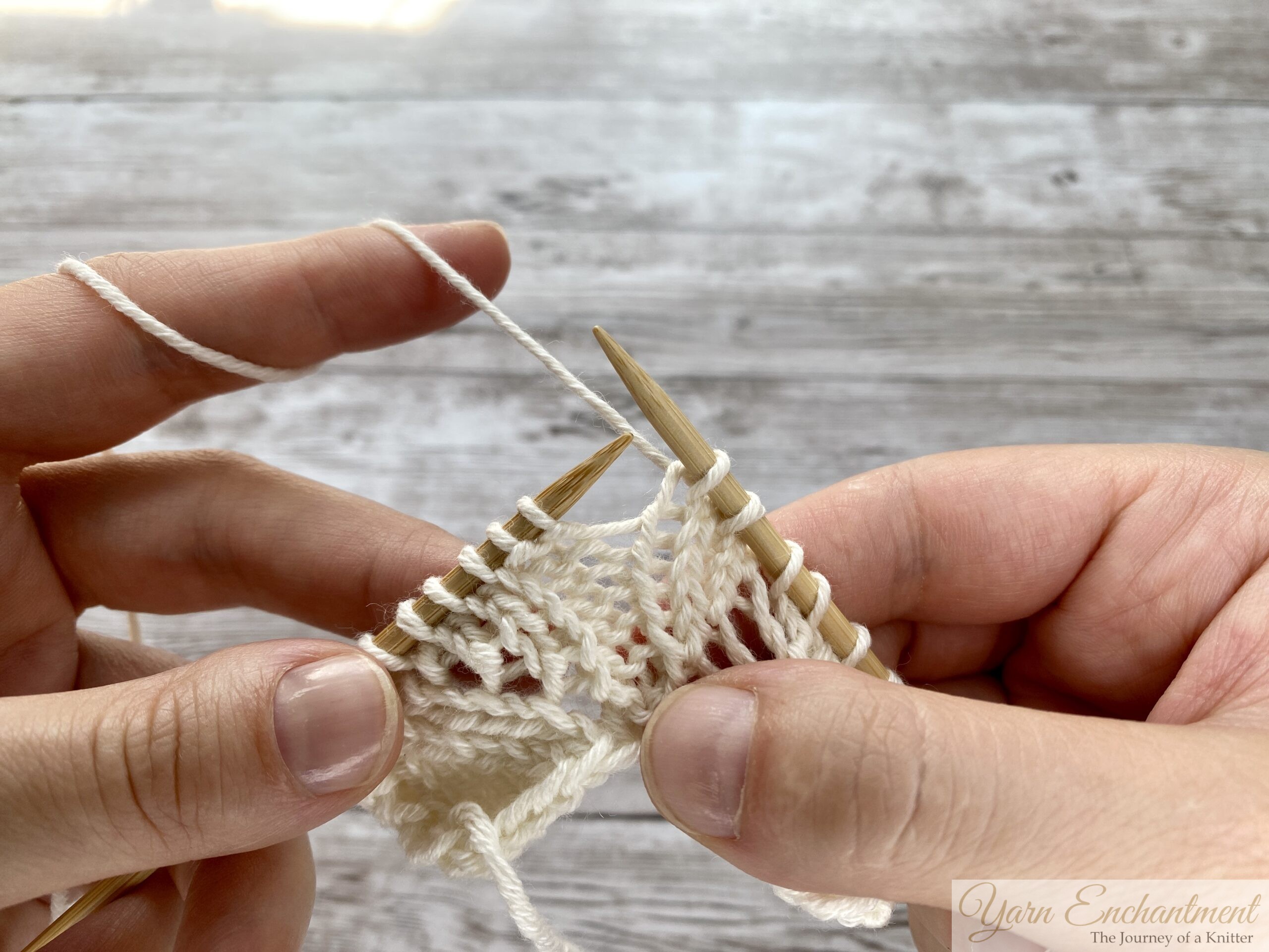 Close-up of hands knitting with wooden needles. The working yarn has been brought over the right needle to create a yarn over loop in cream lace knitting.