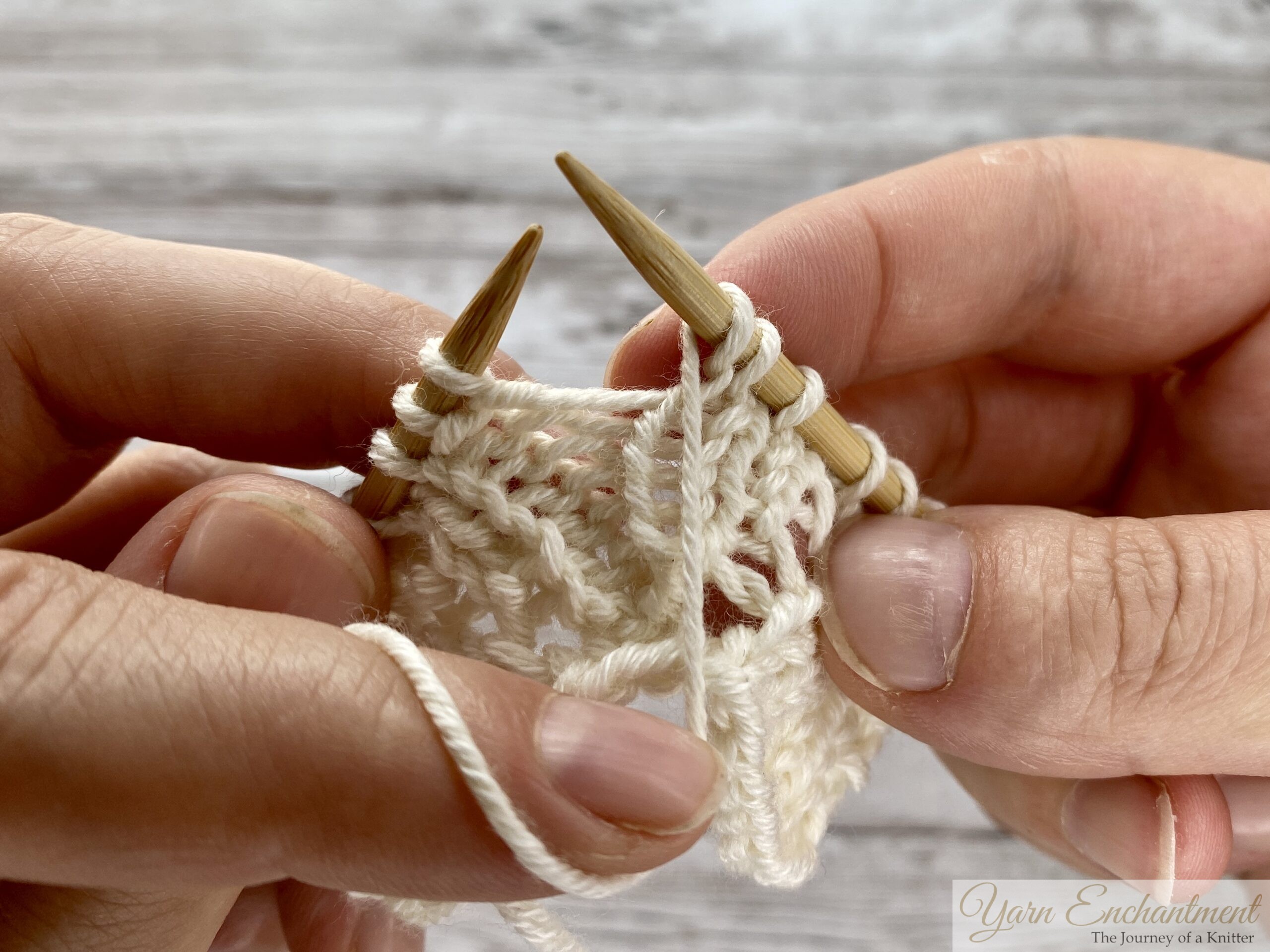 Close-up of hands knitting. The right needle, with the yarn over loop still on it, is pushed into the next stitch on the left needle, preparing to pull the yarn through.