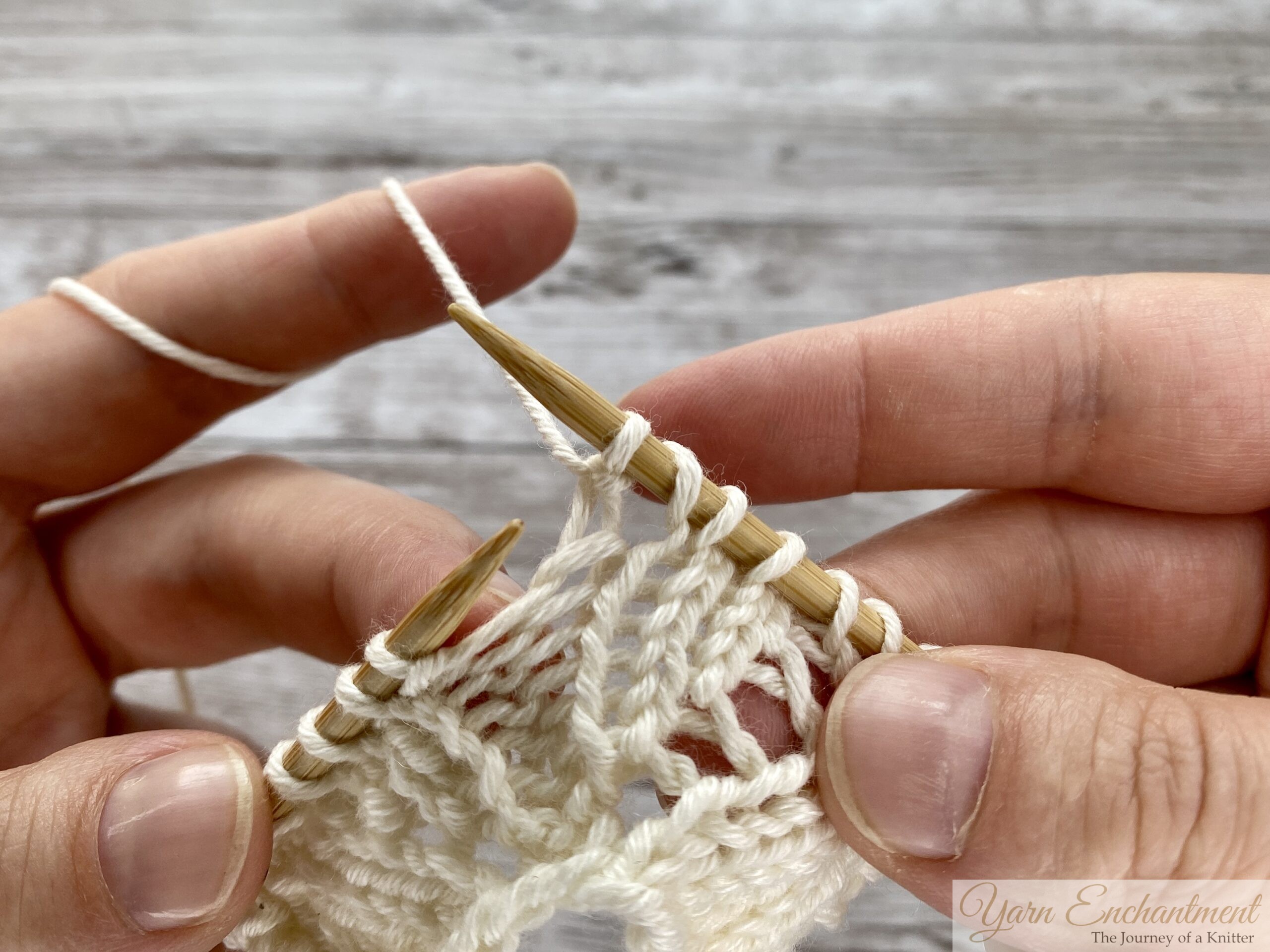 Close-up of hands finishing a lace stitch. The new stitch has been pulled through after the yarn over, securing the loop and creating the open, lacy hole in the cream fabric.