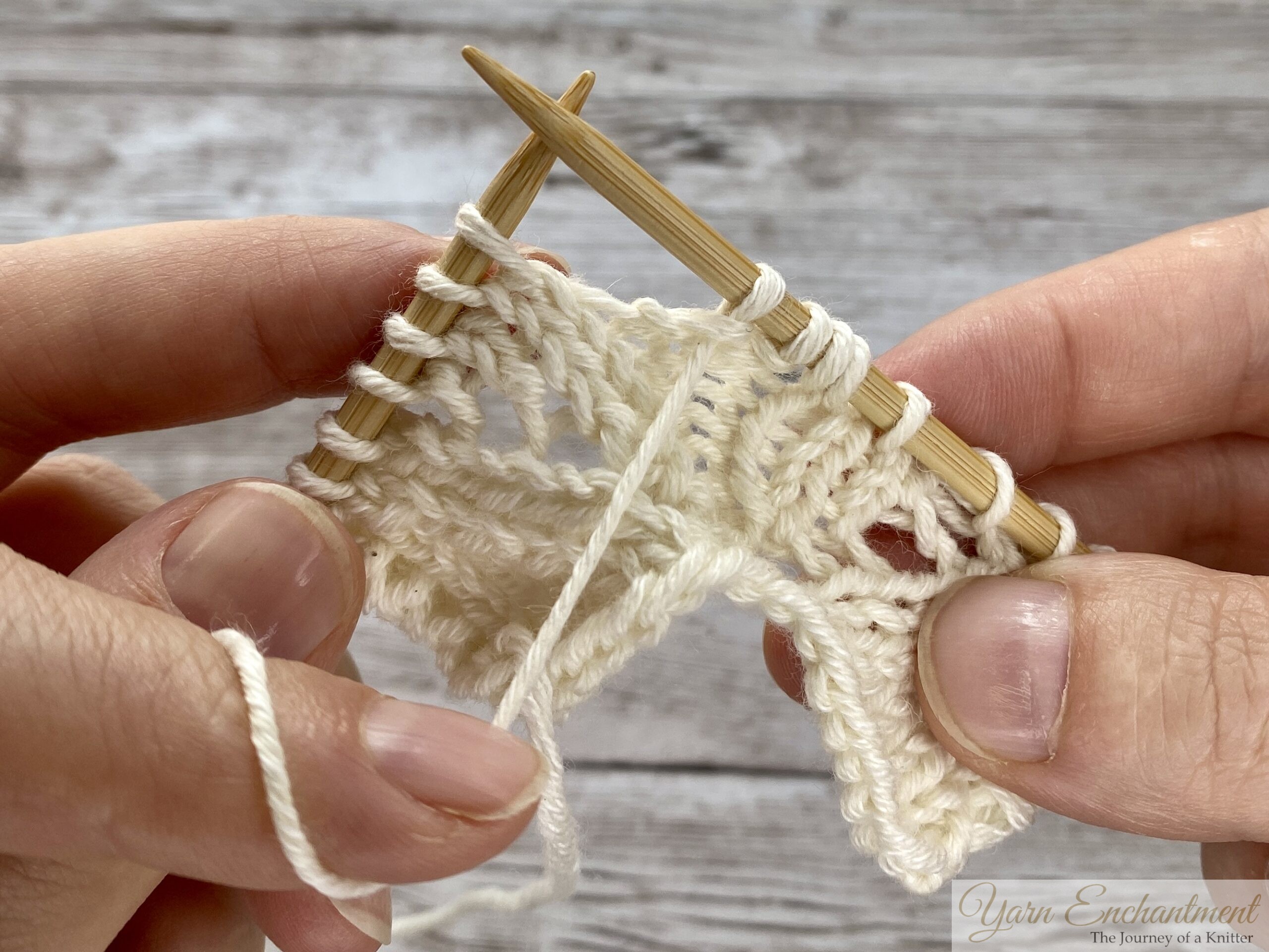 Close-up of hands knitting lace with wooden needles. The working yarn is in the front, having just completed a purl stitch, and the right needle is ready to be moved to create a yarn over before the next knit stitch.