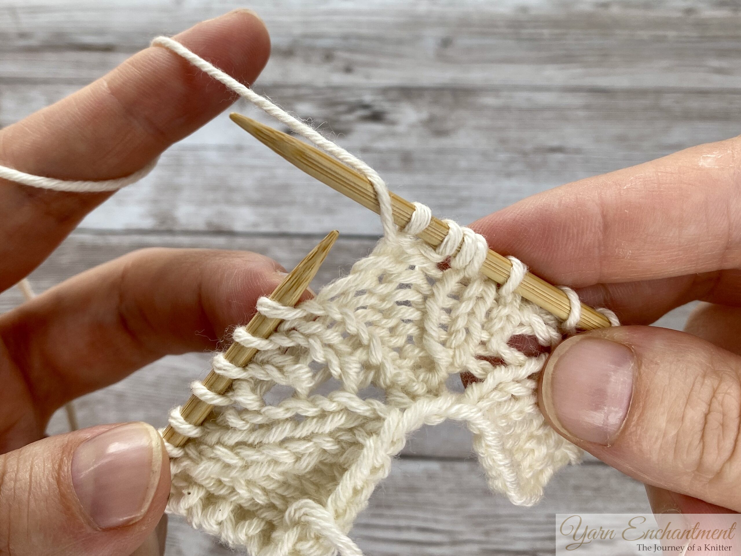 Close-up of hands knitting. The yarn is being lifted from the front to the back over the right needle to create a yarn over loop, transitioning between a purl and a knit stitch in cream lace knitting.