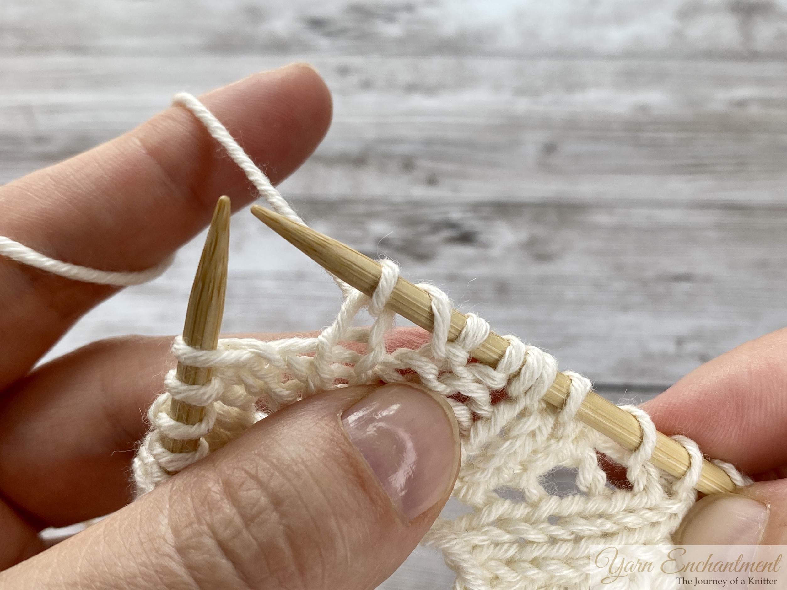 Close-up of hands completing the yarn over. The new loop is secured on the right needle, and the needle is now positioned to insert into the next stitch to complete a knit, creating a lacy hole in the fabric.