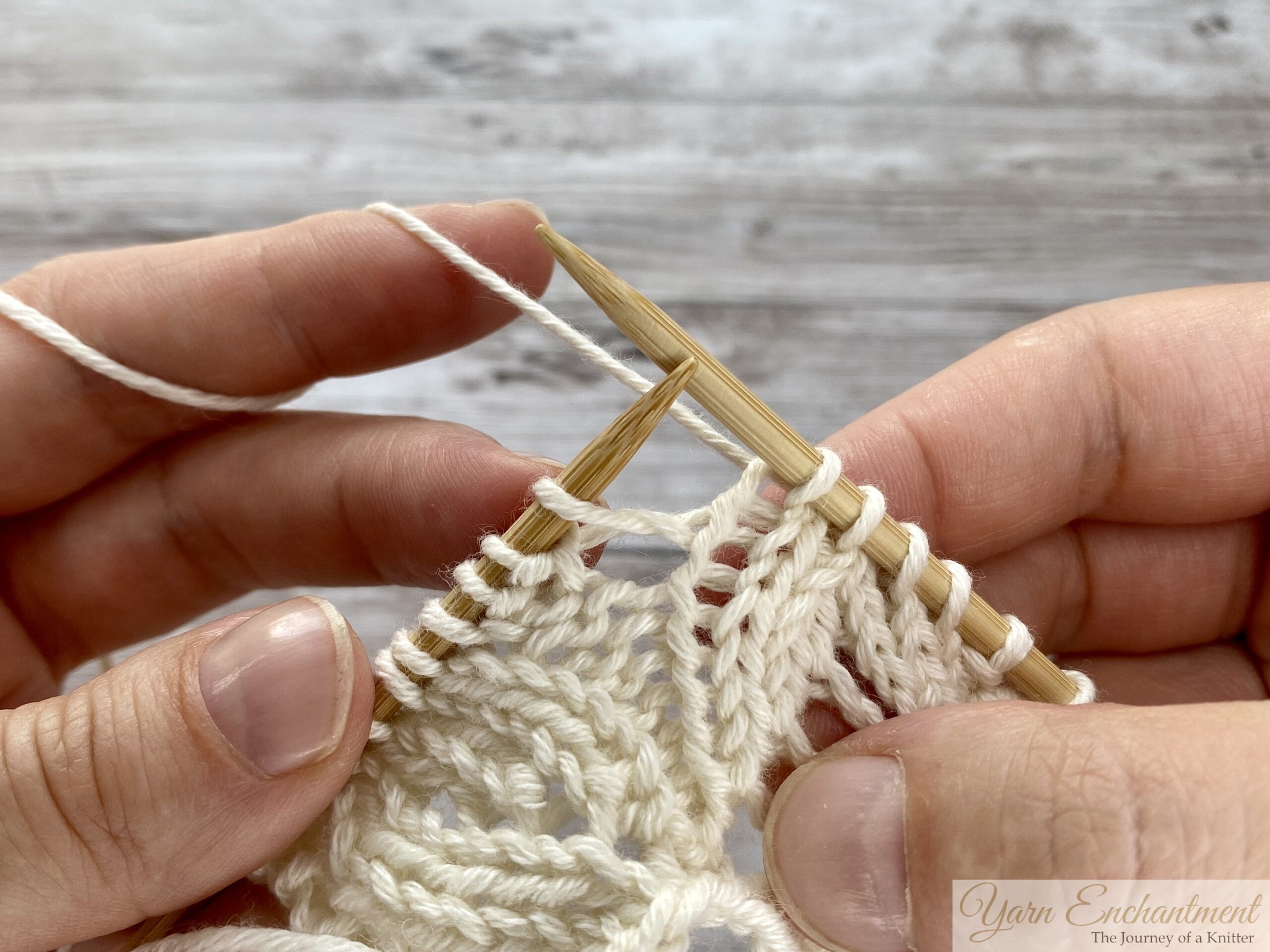 Close-up of hands holding wooden knitting needles mid-row. The right needle is poised to enter the next stitch on the left needle, preparing to work a yarn over between two knit stitches in cream lace knitting.