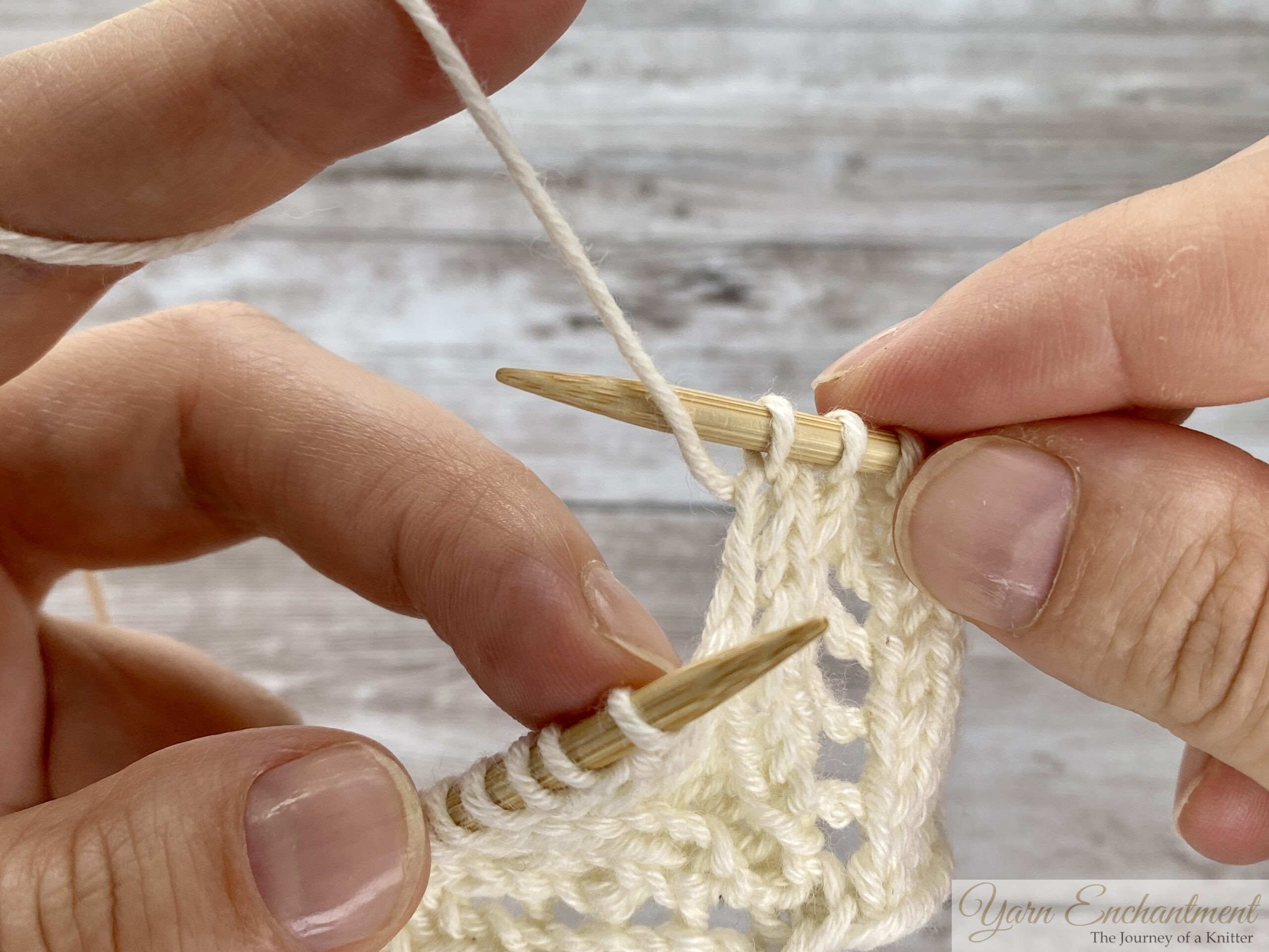 Close-up of hands knitting. The right needle is inserted into the next stitch while the working yarn is being lifted from the front over the right needle, creating the yarn over loop in lace knitting.