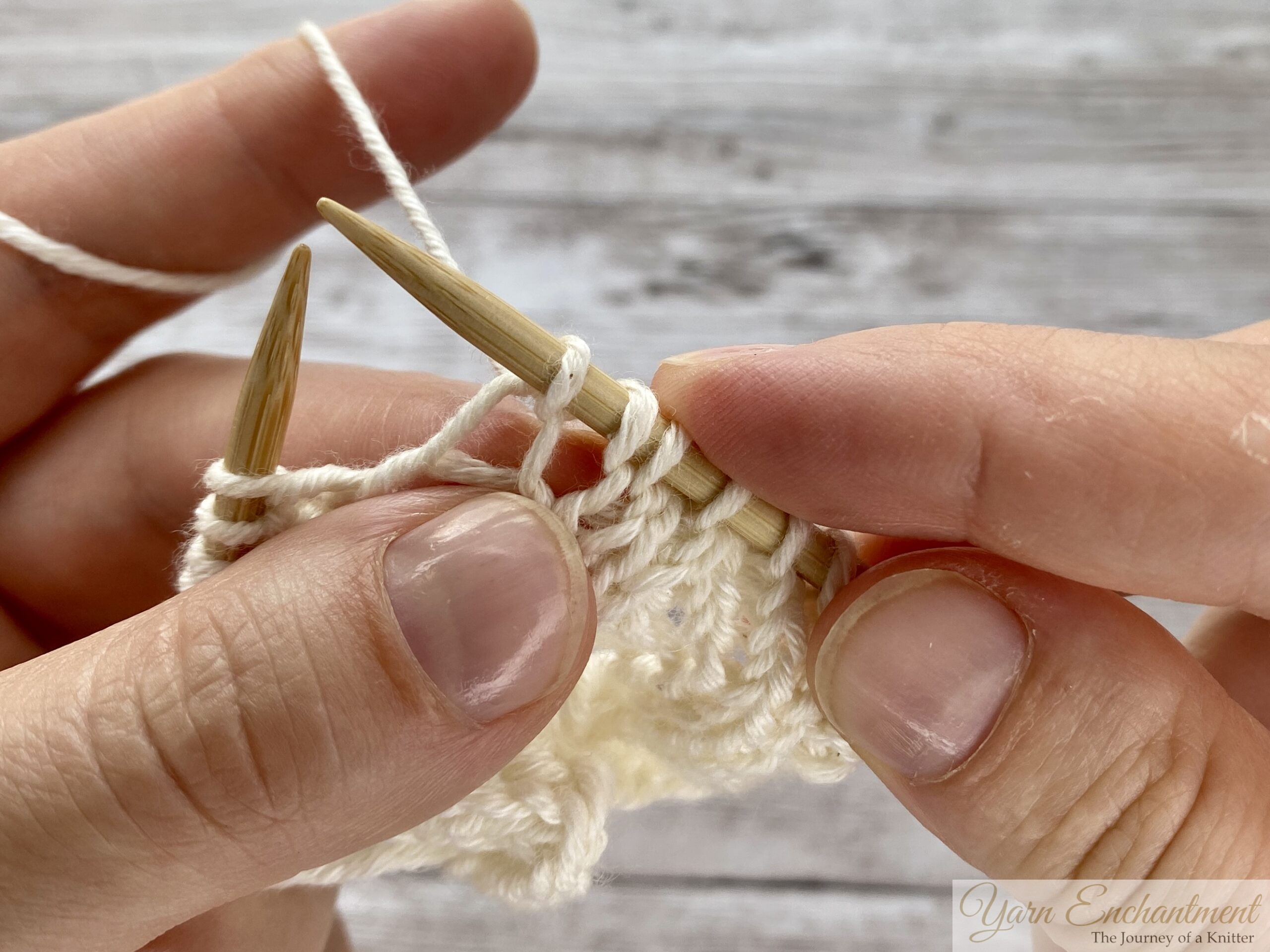 Close-up of hands completing the sequence. The right needle is pulling the new stitch through after the yarn over, securing the new open loop into the lace pattern.