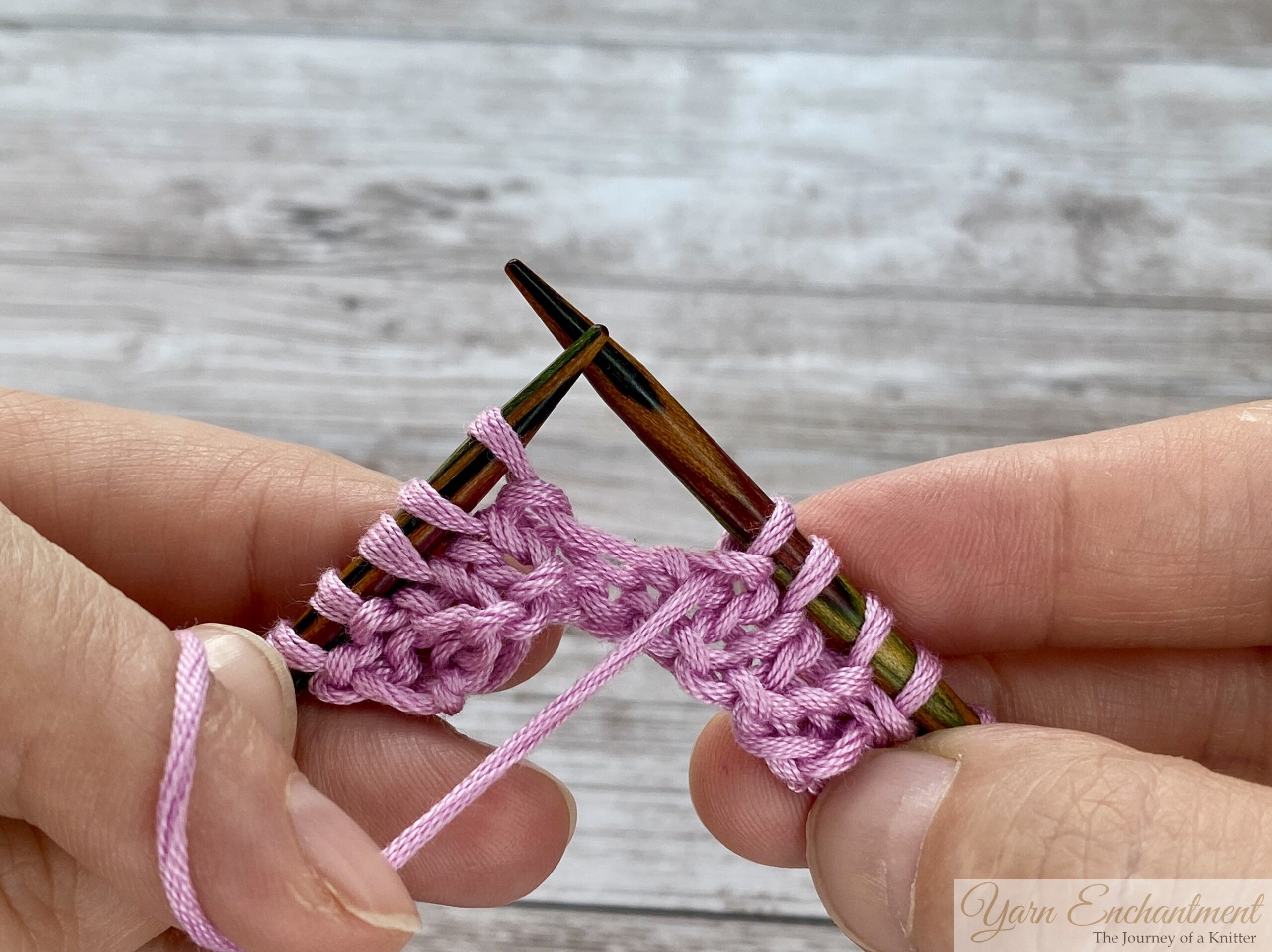 Close-up of hands knitting with dark wooden needles and pink yarn. The working yarn is in the front, having just completed a purl stitch, preparing to create a yarn over before the next purl stitch.