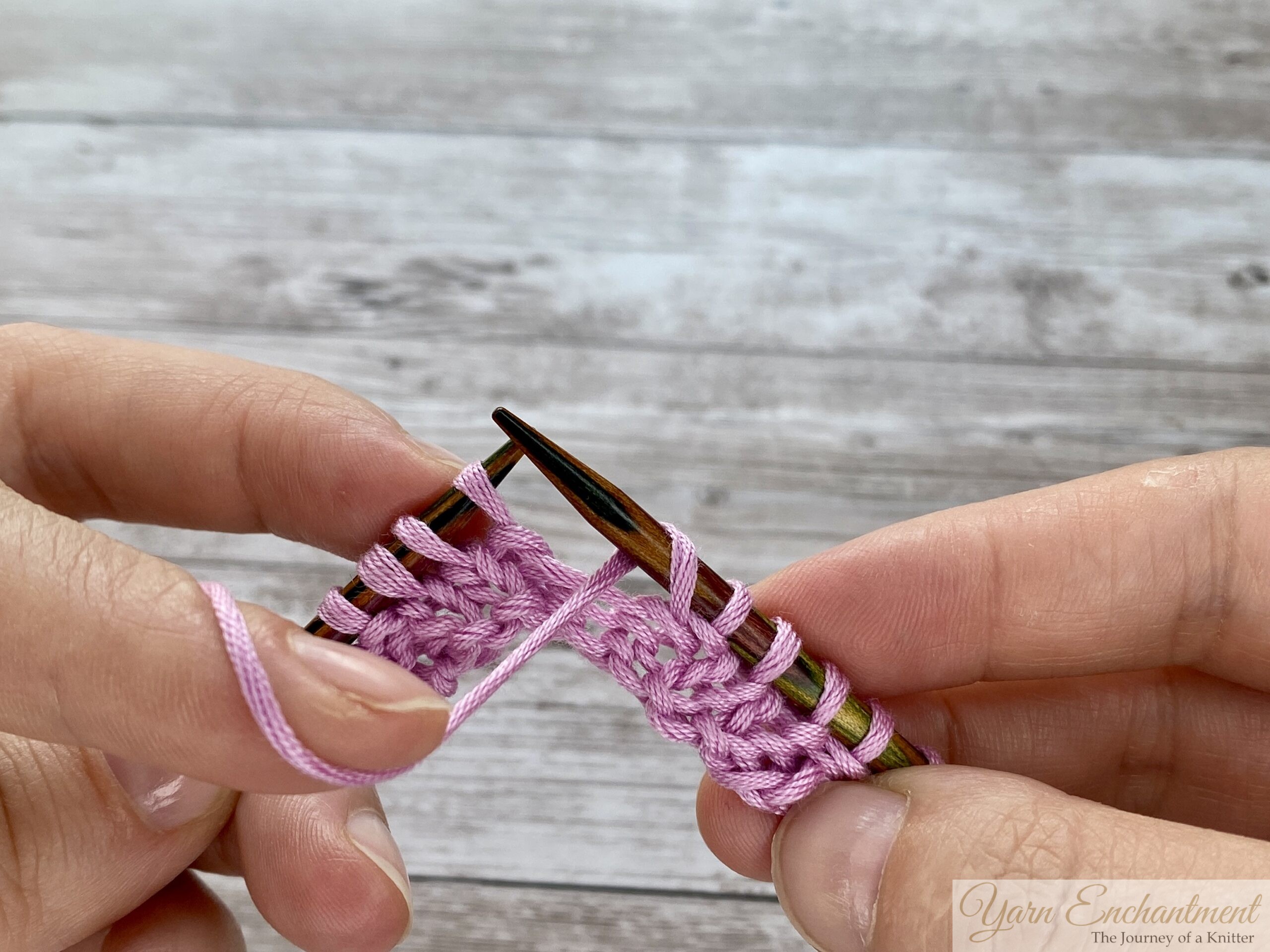 Close-up of hands completing the yarn over. The new loop is fully formed and resting on the right needle, and the working yarn is in the front, ready to be purled into the next stitch on the left needle.