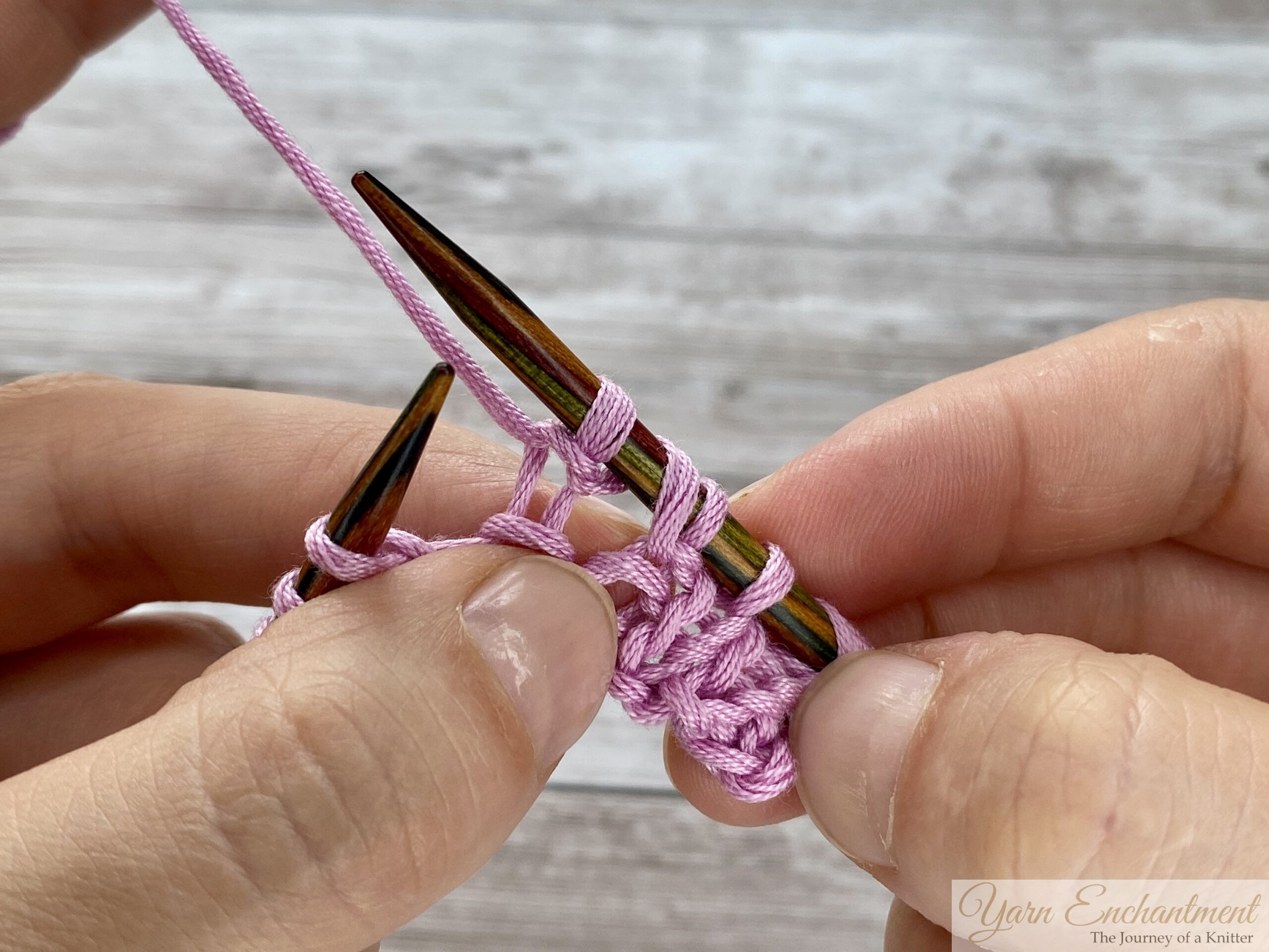 Close-up of hands knitting lace. The working yarn is being lifted from the front, wrapped over the right needle to the back, and is coming back to the front to create the yarn over loop between two purl stitches.