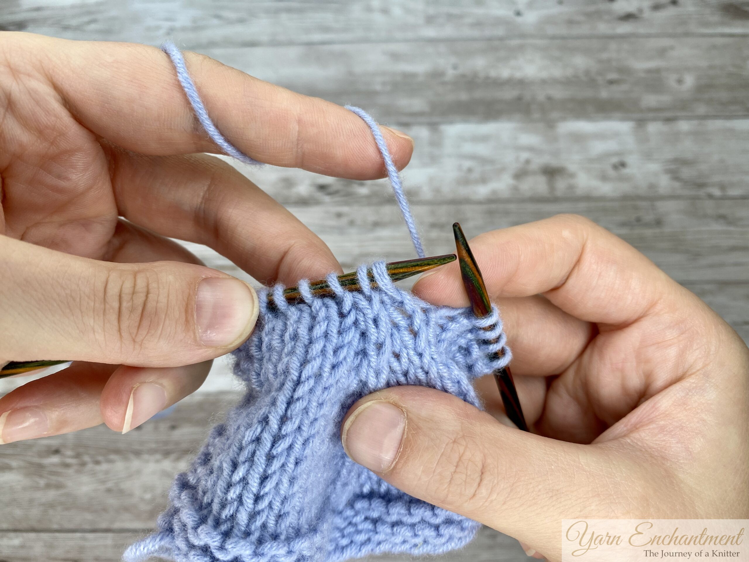 A close-up of a knitter's hands holding wooden needles and light blue yarn, demonstrating the first step of a knitting technique against a rustic wooden background. The knitter has inserted the right needle tip into three stitches on the left needle simultaneously, preparing to slip them together as if to knit.