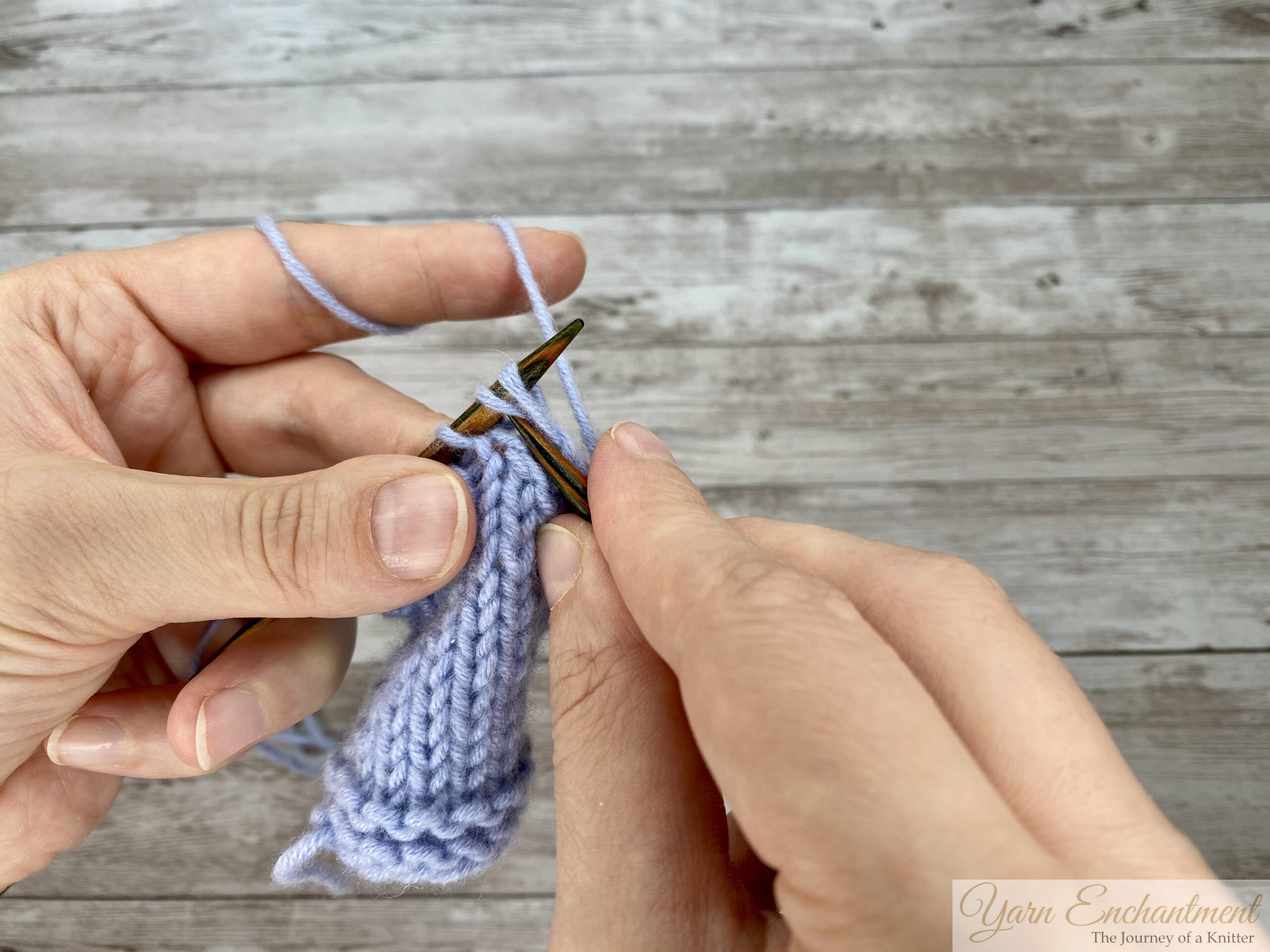 A close-up of knitter's hands working light blue yarn on wooden needles, showing the middle step of a knitting maneuver. The right needle tip is being used to lift the three slipped stitches back over the newly knitted stitch from the previous step, effectively combining them.