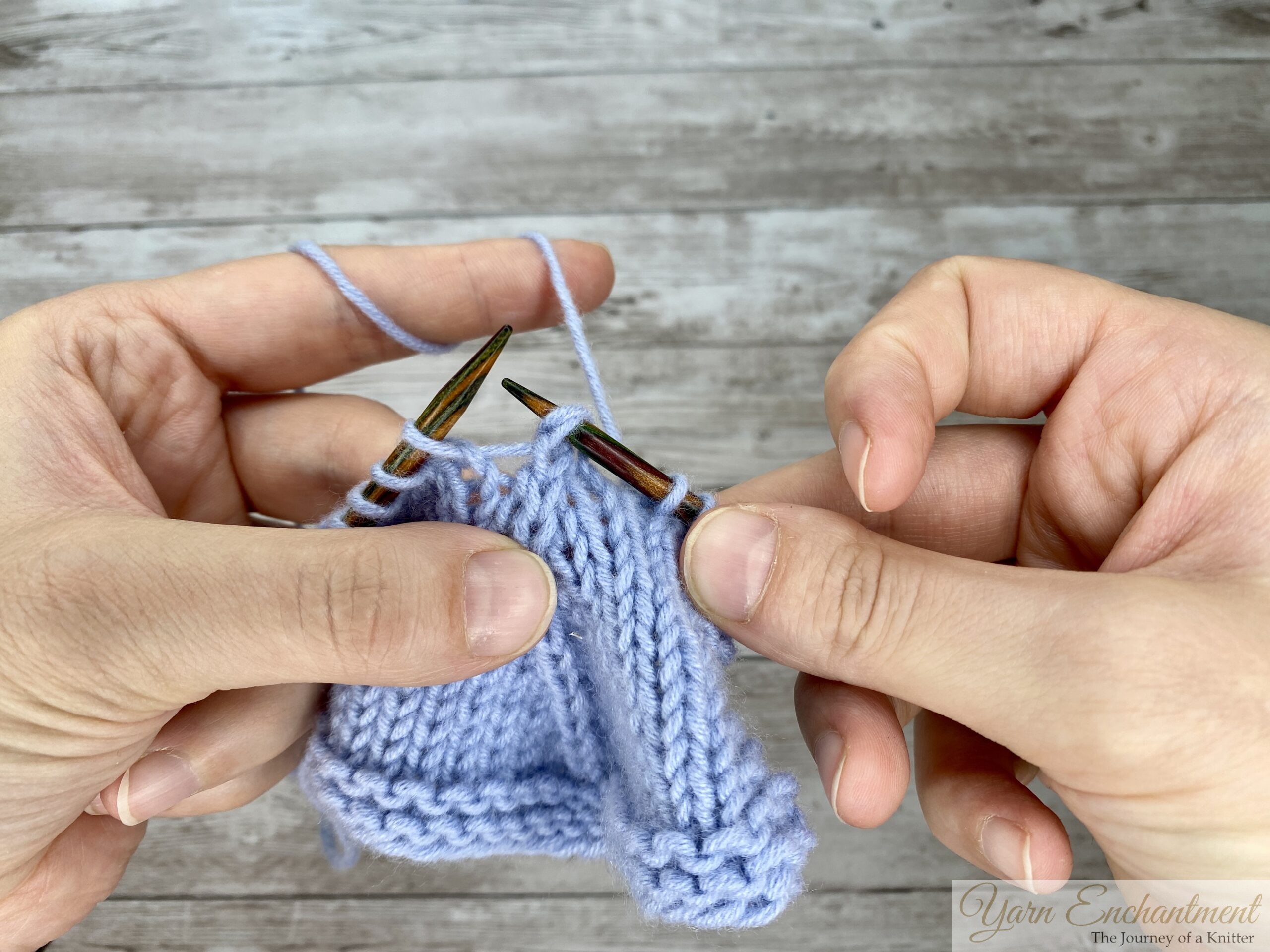 A close-up of knitter's hands showing the completed stitch of a knitting technique on a light blue swatch against a wooden background. The right needle holds the single, finished stitch that was created by reducing three original stitches down to one, which sits at the peak of a visible, centered raised line on the knitted fabric below.