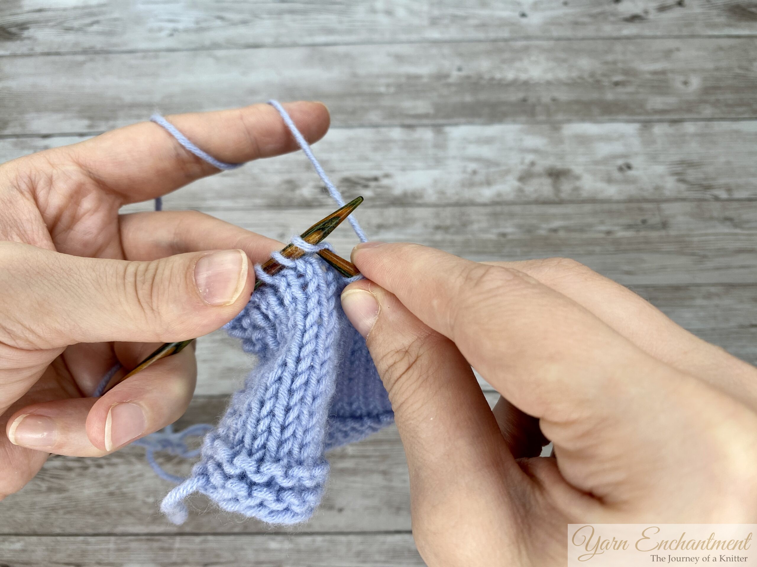 A close-up view of a knitter's hands demonstrating a stitch on a light blue knitted swatch against a weathered wooden background. The knitter is holding the working yarn taut with their left hand, while the right hand uses the tip of the right wooden knitting needle to lift a stitch over another stitch on the needle, completing a decrease maneuver.