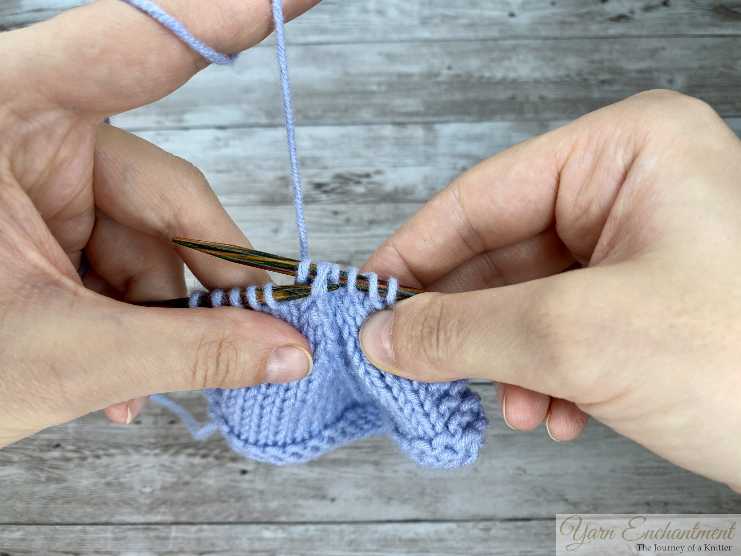 A close-up of knitter's hands working on a light blue swatch with wooden needles against a wooden surface. The knitter is in the middle of a knitting action, holding the fabric firmly with both hands as the right needle interacts with the stitches on the left needle, with the working yarn tensioned over the left index finger.