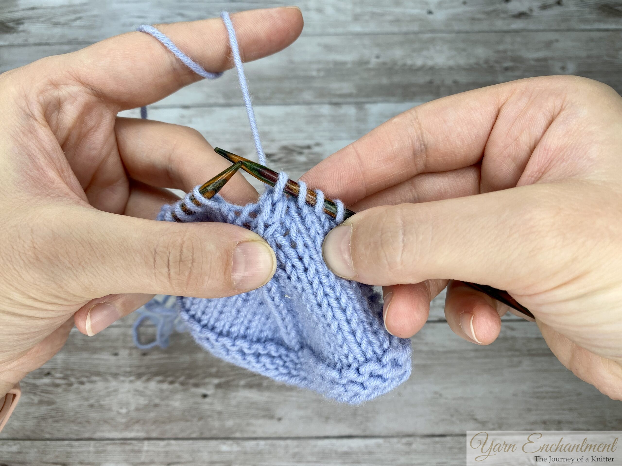 A close-up of a knitter's hands holding wooden needles and light blue yarn over a wooden background, demonstrating a stitch. The right needle has been inserted into a stitch on the left needle in a way that suggests the start of a slipping maneuver, with the working yarn held up by the left index finger.