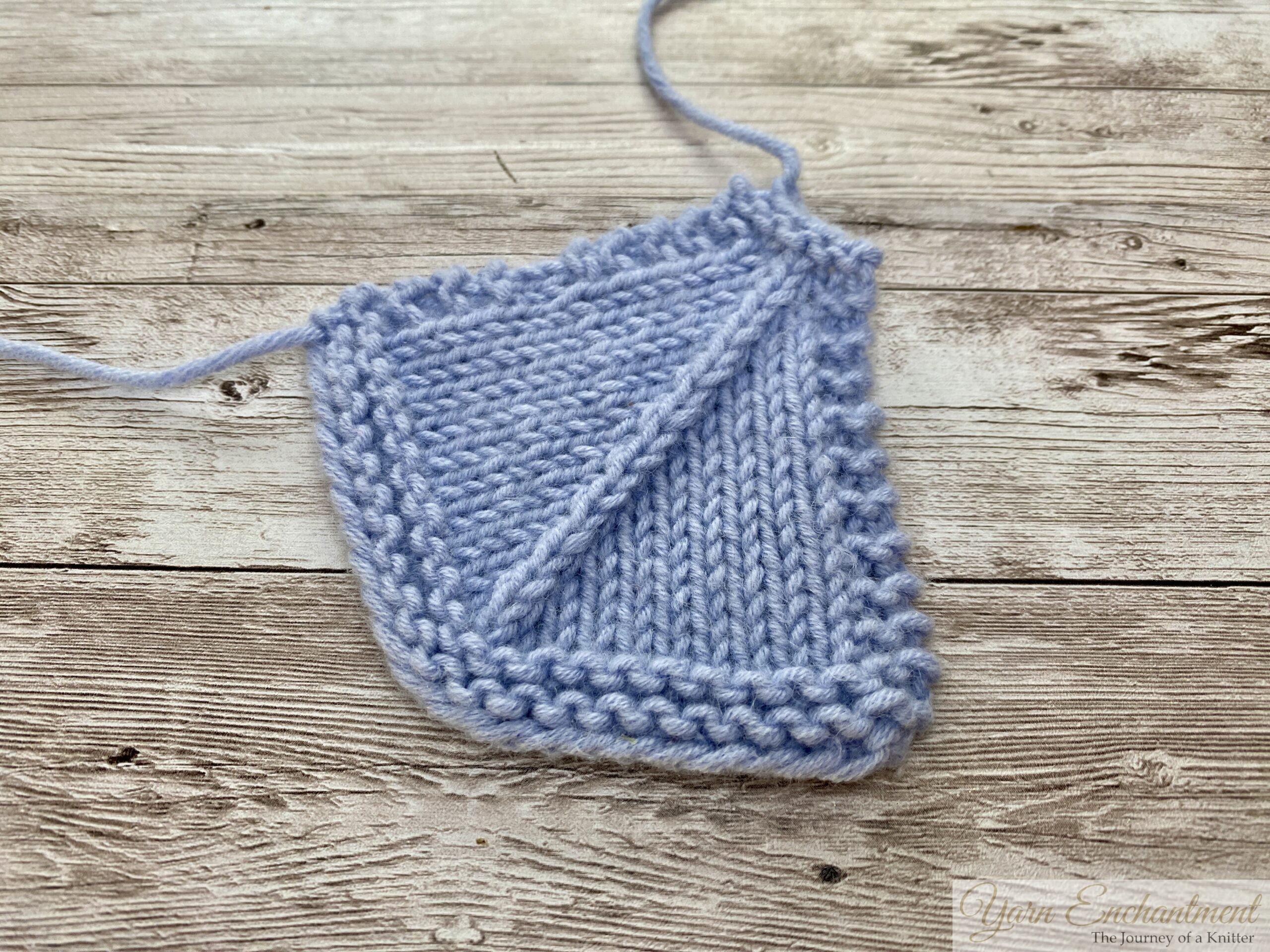 A light blue knitted swatch resting on a wooden surface, demonstrating a 'Centered Double Decrease' in knitting. The stitch pattern shows a distinct, straight diagonal line moving upwards from left to right through the center of the stockinette fabric, bordered by garter stitch edges.