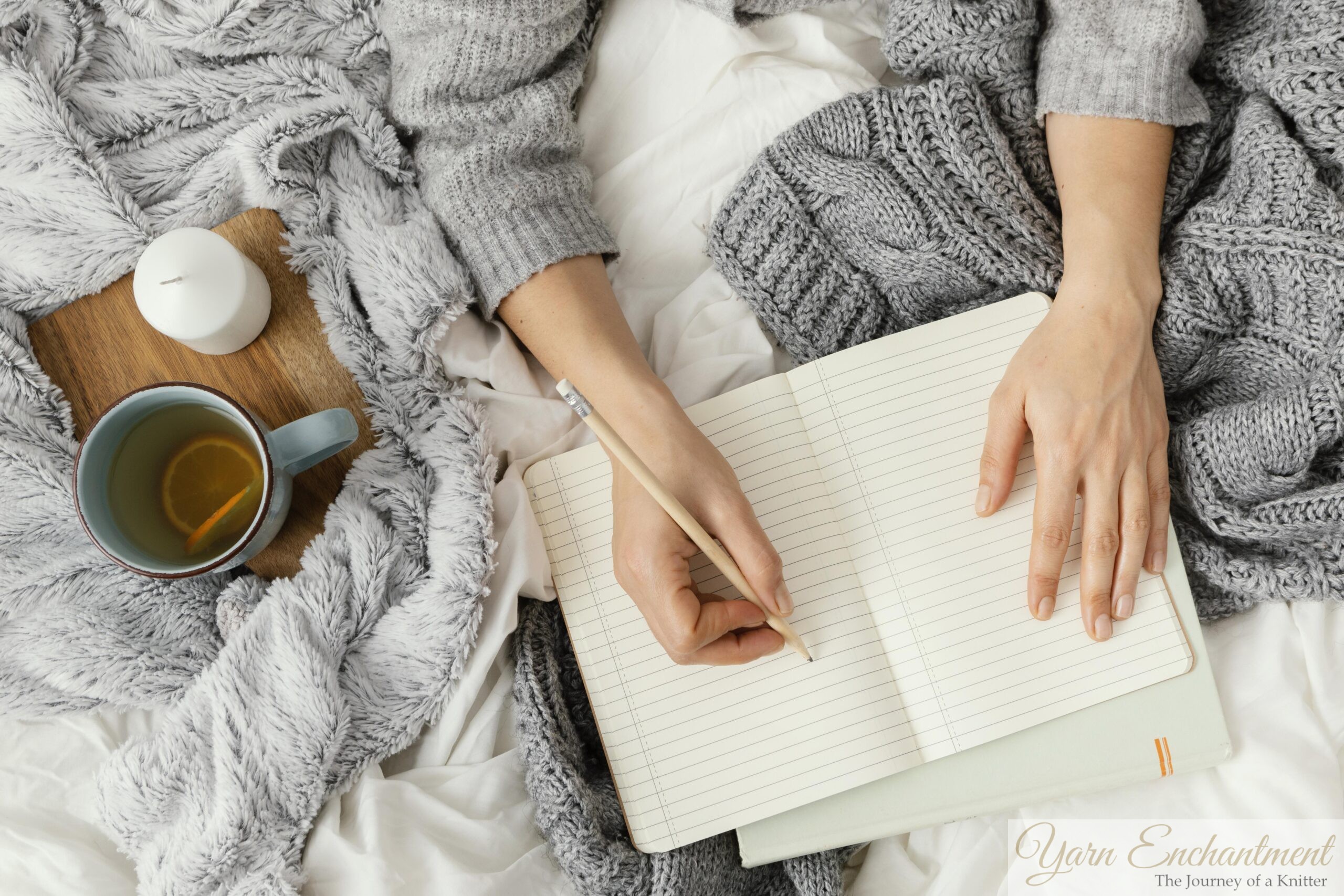 A top-down flat lay of hands writing in an open lined journal with a wooden pencil, next to a warm mug of lemon tea and a white candle, resting on soft white bedsheets with a grey knitted sweater and plush blanket.