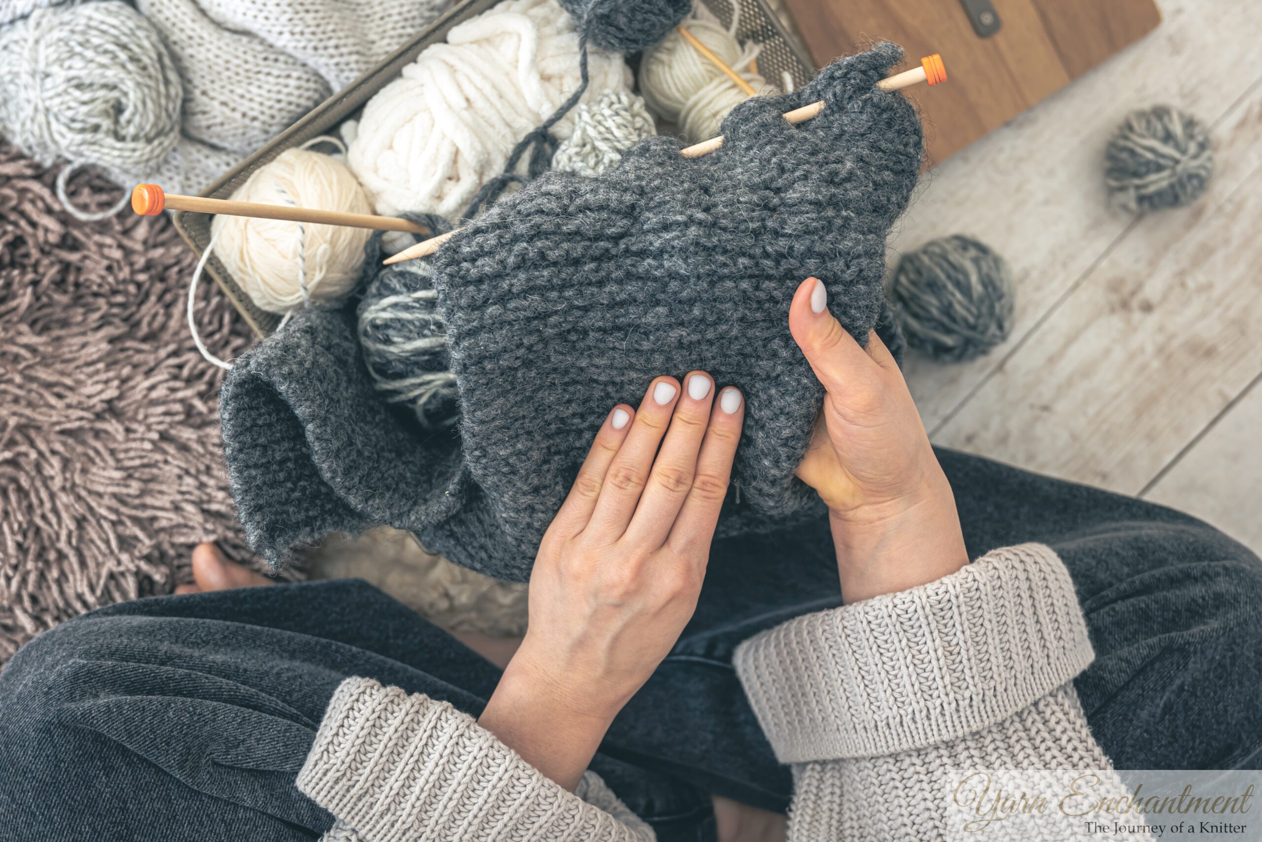 A first-person view of hands gently resting on a dark grey knitted scarf, paused mid-stitch in a quiet moment of