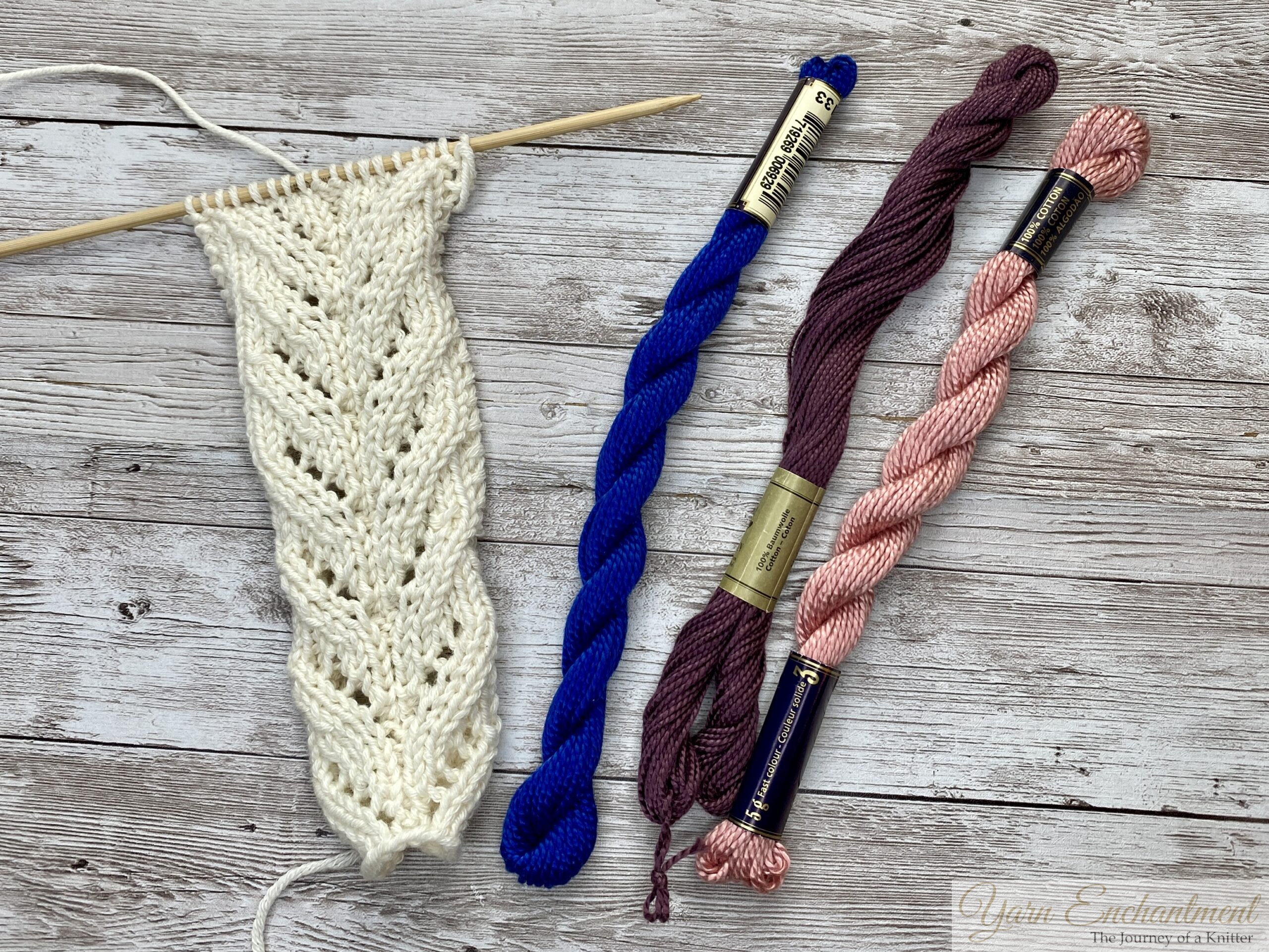 Cream lace swatch on a bamboo needle beside three cotton skeins in blue, plum, and pink, lifeline thread options for lace knitting.