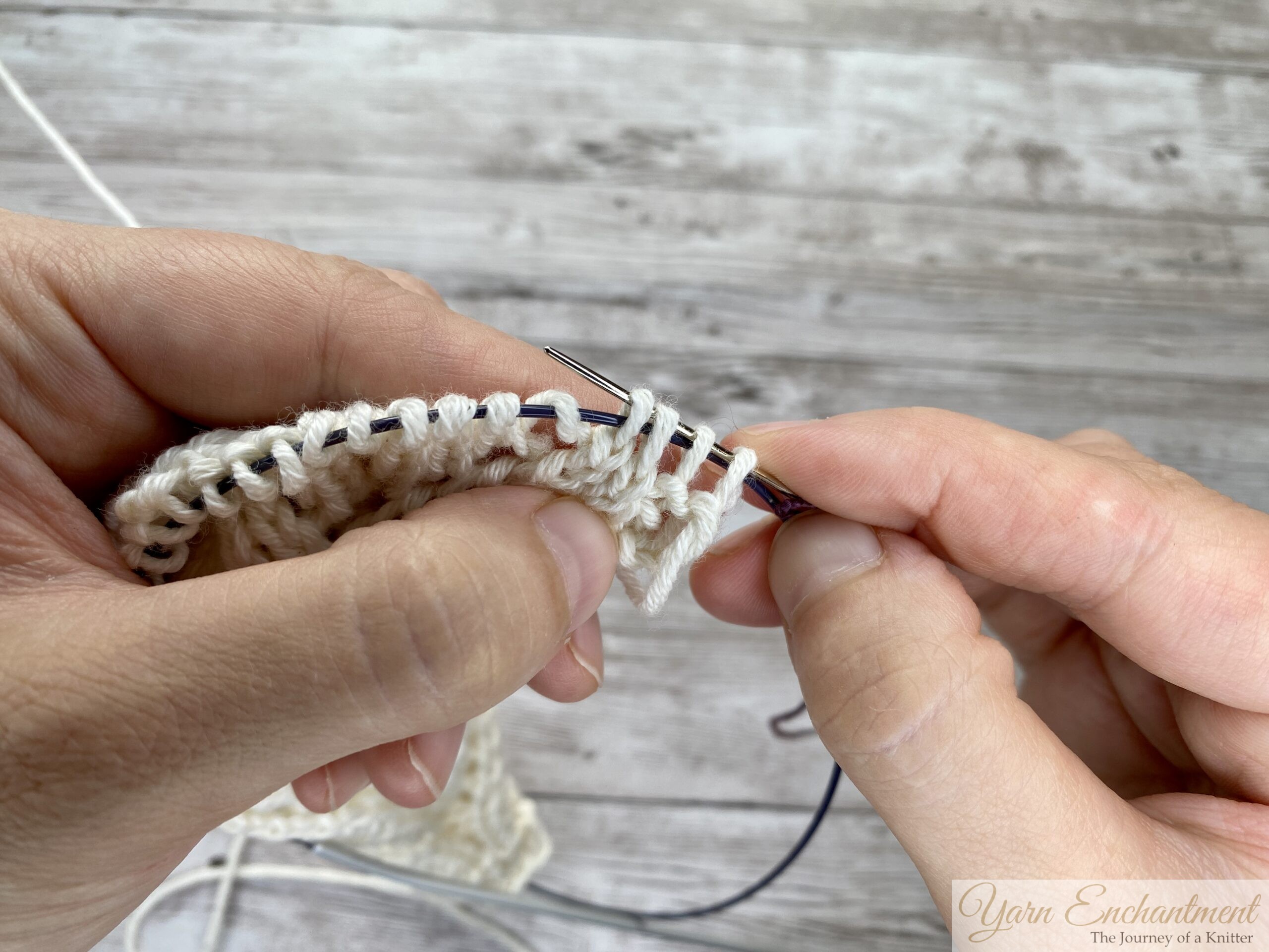 Tapestry needle sliding through several stitch loops on the circular needle cable, plum lifeline thread following behind.